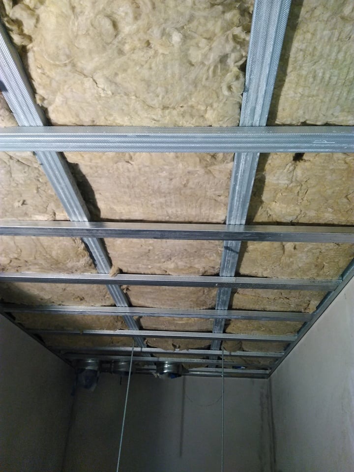 Sufit podwieszany w trakcie montażu: metalowa konstrukcja z widoczną izolacją z wełny mineralnej, przewody wentylacyjne wystające z sufitu.