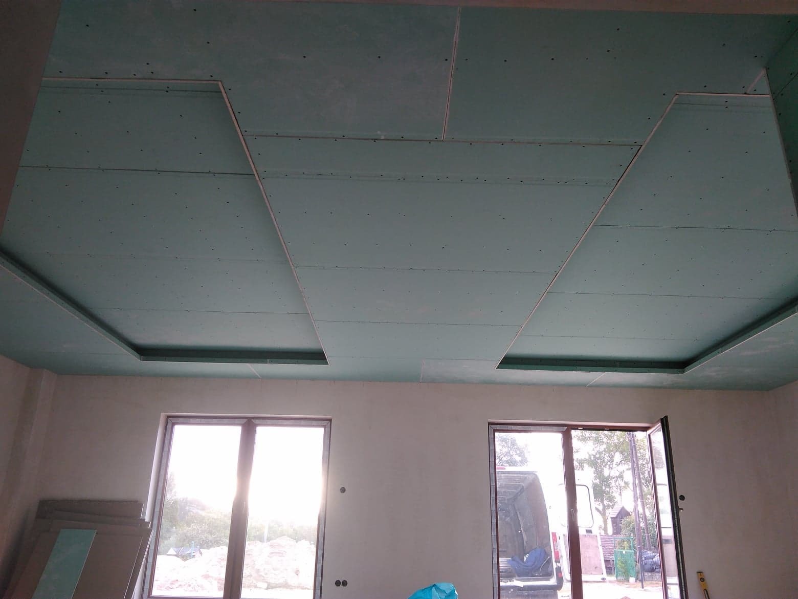 Montaż sufitu podwieszanego z płyt gipsowo-kartonowych z widocznymi otworami na oświetlenie w nowym pomieszczeniu z dwoma oknami.