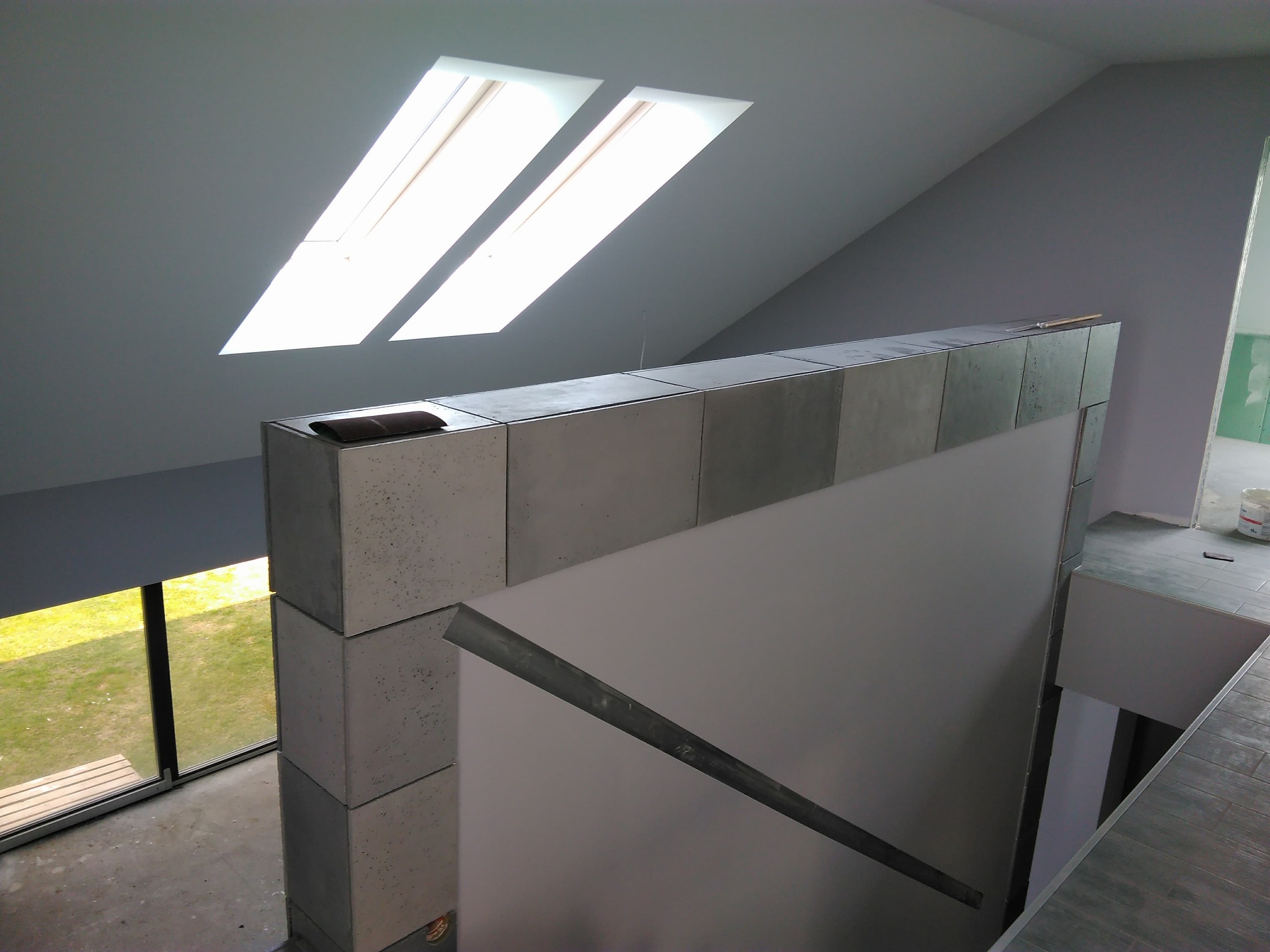 Wnętrze domu w trakcie wykańczania: betonowe bloczki tworzą ściankę działową pod skosem dachu z oknami, widoczne surowe ściany i podłoga w szarych płytkach, w tle fragment pomieszczenia w trakcie...