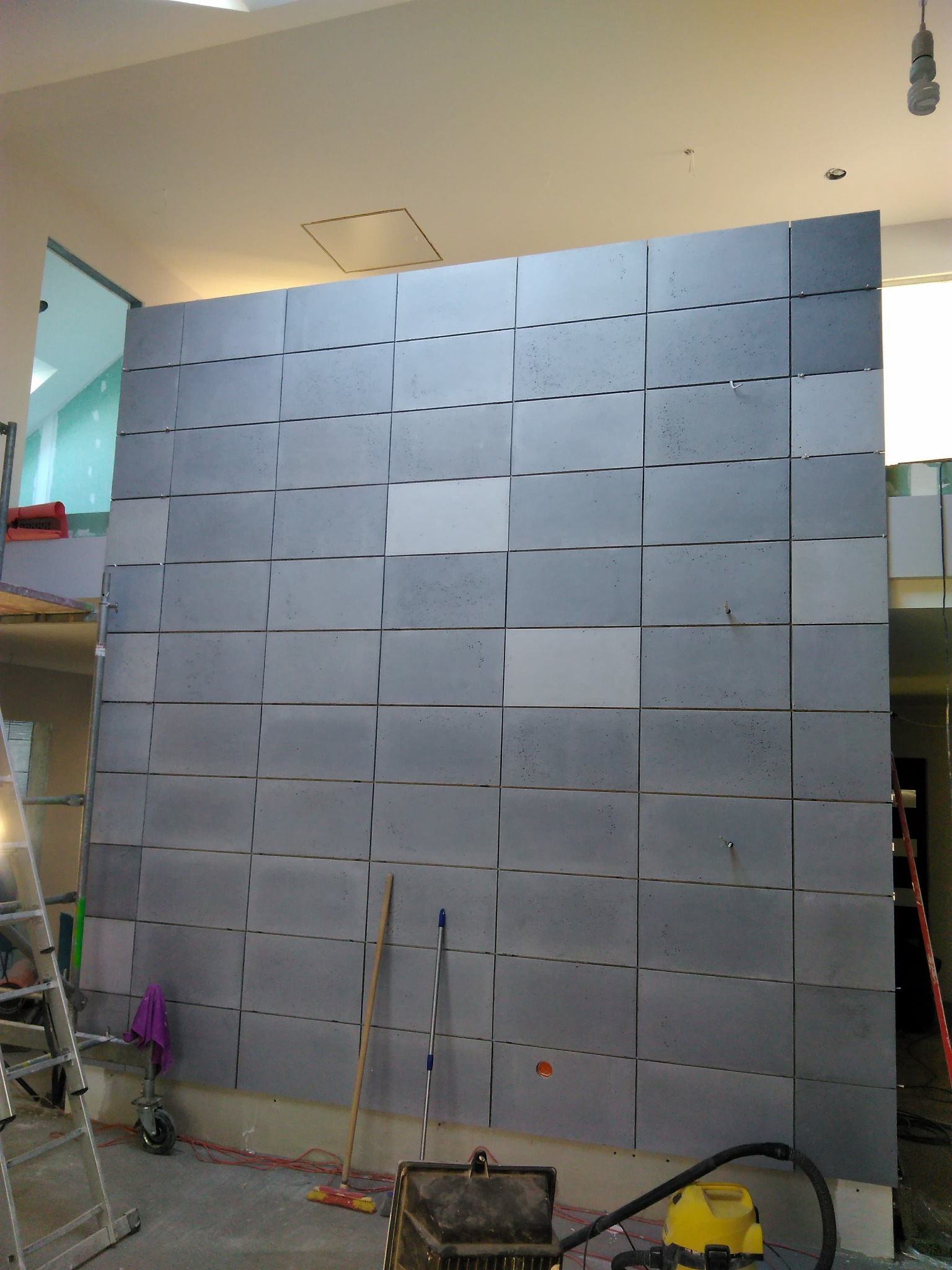 Montaż szarych, betonowych płyt na ścianie w Dychowie, widoczne narzędzia i drabina, etap wykończeniowy.