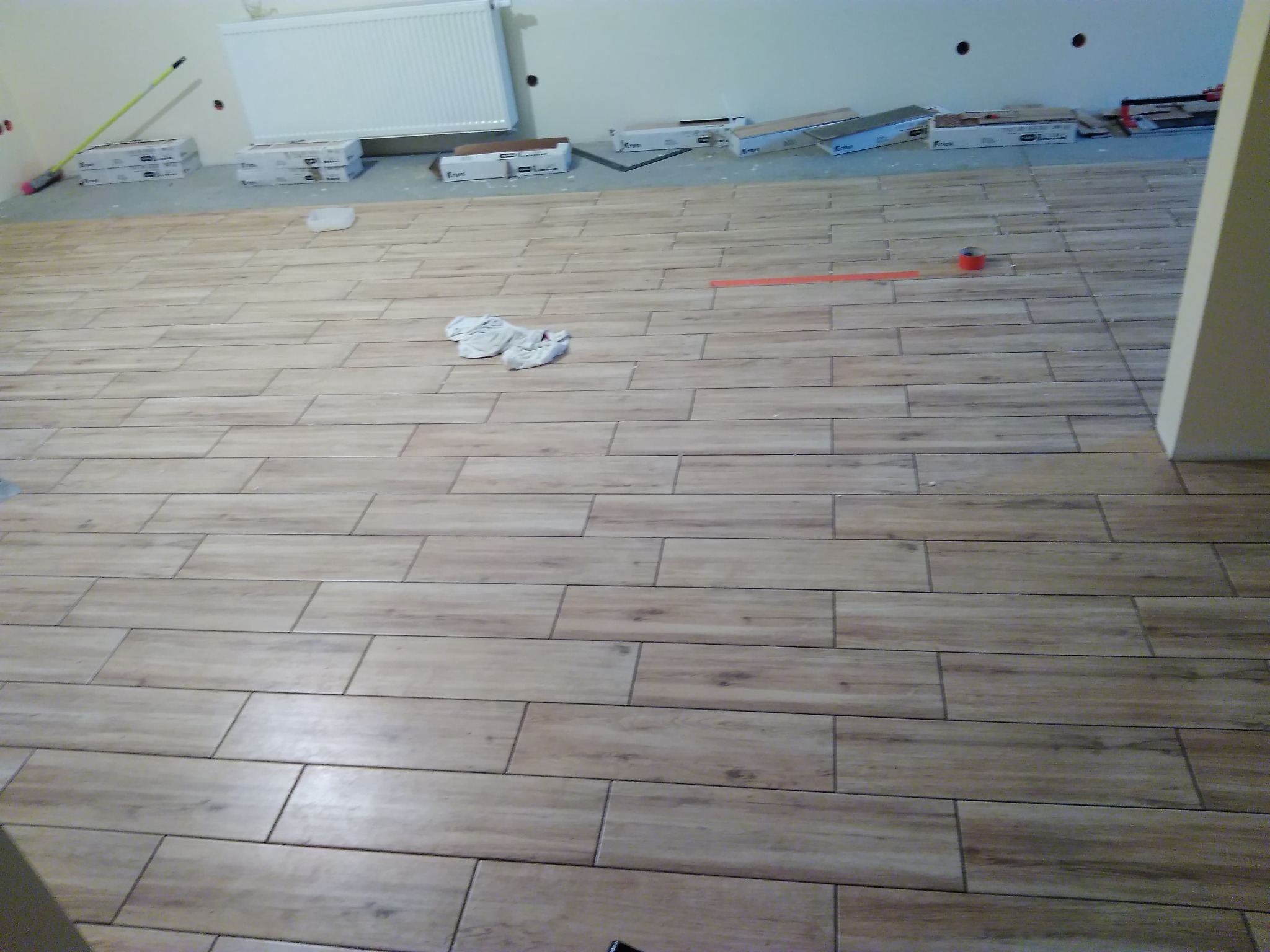 Ułożona podłoga z płytek imitujących drewno, widoczne pudełka po płytkach przy ścianie, pomarańczowa miarka i czerwona taśma malarska, oraz białe rękawice robocze.