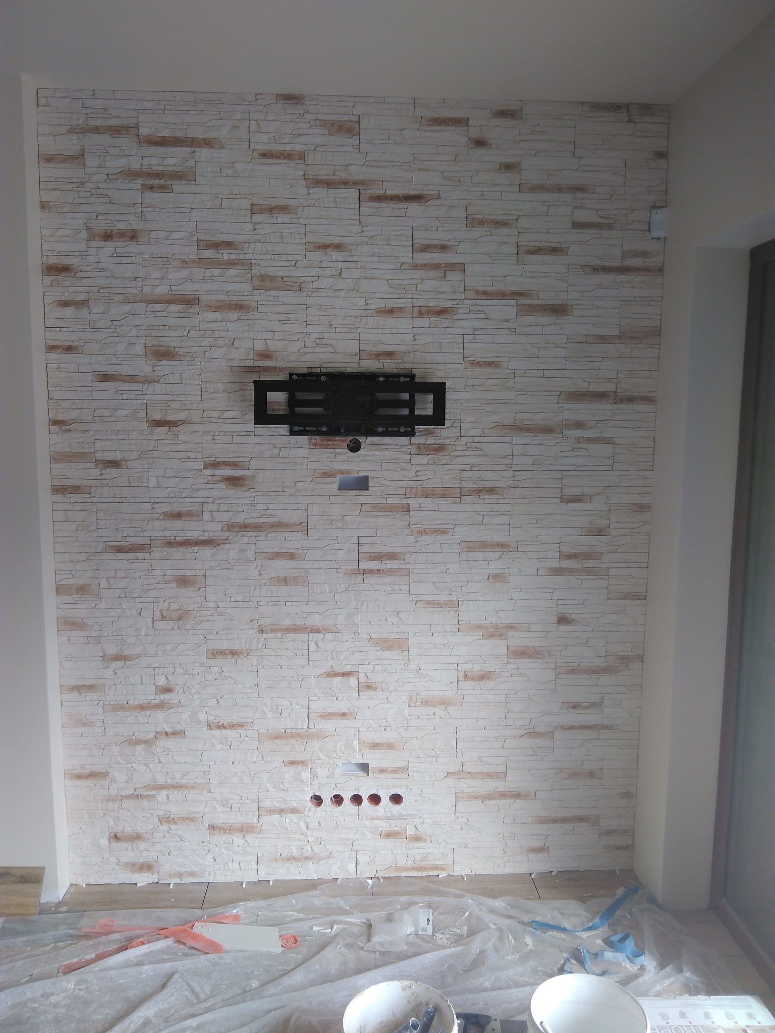 Ściana wyłożona dekoracyjną cegłą w odcieniach bieli i beżu, z zamontowanym uchwytem do telewizora i przygotowanymi otworami na przewody, zabezpieczona folią malarską podczas prac wykończeniowych.