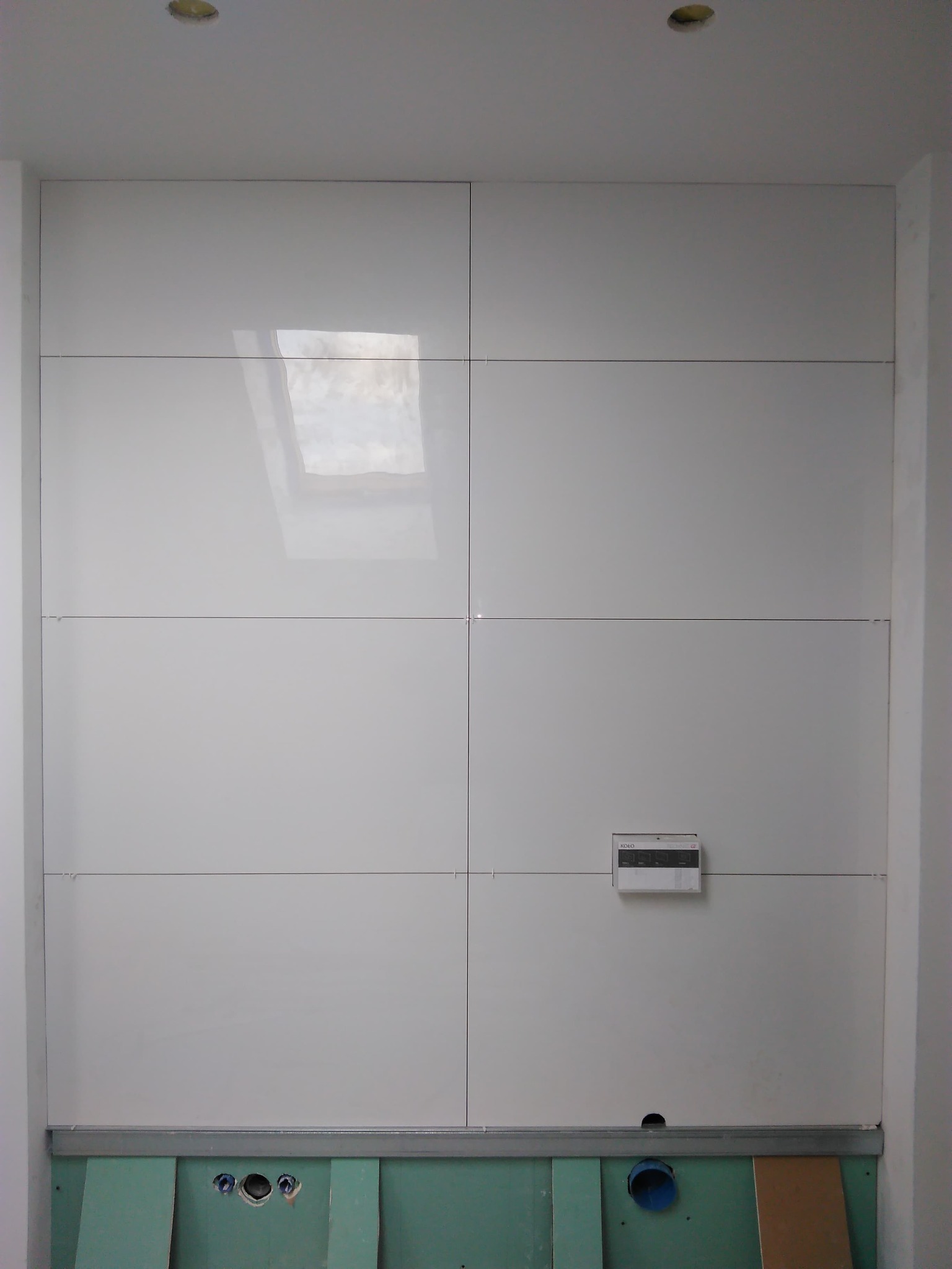 Ściana wyłożona białymi, błyszczącymi panelami z widocznym panelem sterowania i przygotowanym miejscem na instalacje, dolna część ściany z zielonymi płytami gipsowymi i otworami na rury.