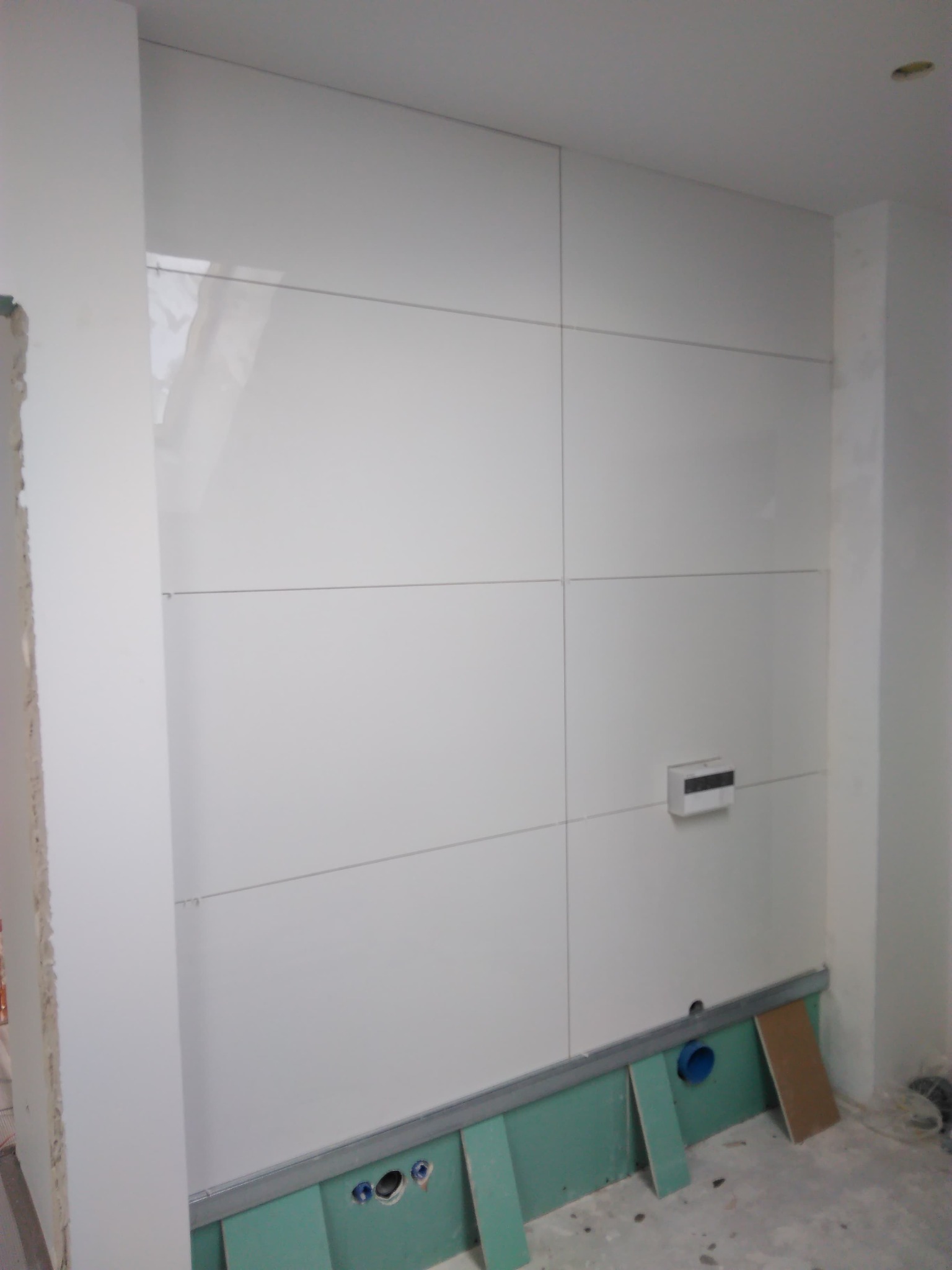 Biała ściana wyłożona błyszczącymi panelami w trakcie montażu, widoczne pionowe i poziome fugi, dolna część ściany z zielonymi płytami gipsowo-kartonowymi i instalacją hydrauliczną.