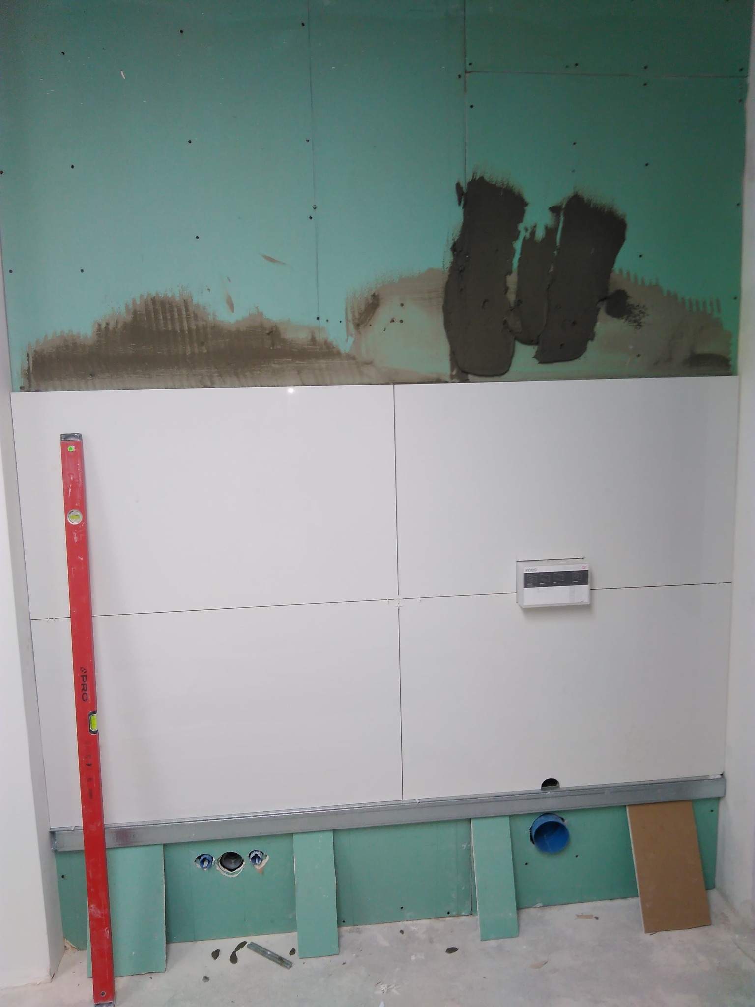 Położone białe płytki na ścianie łazienki z widoczną poziomicą, fragmentem płyty kartonowo-gipsowej oraz przygotowanym miejscem na armaturę.