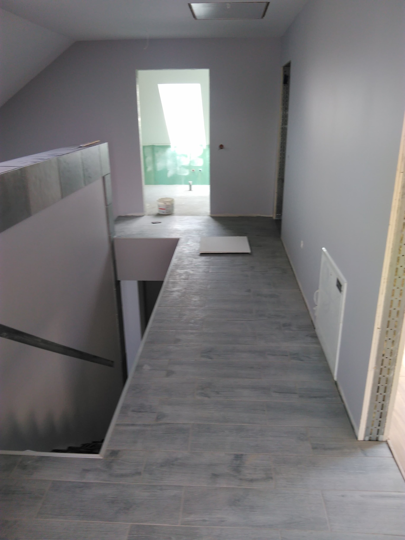 Korytarz w trakcie wykańczania z szarą podłogą imitującą drewno, świeżo pomalowane szare ściany, widoczne wejście do łazienki z wiadrem i niezabezpieczonym oknem dachowym oraz zejście schodami w dół.