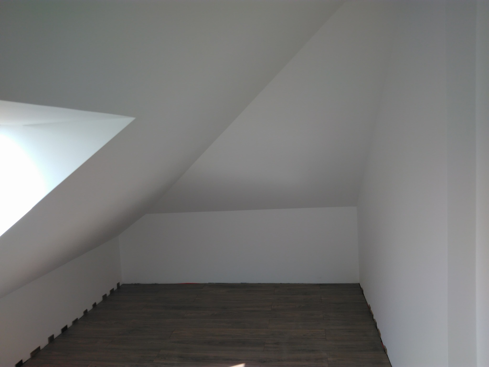 Poddasze po remoncie z nową podłogą z paneli o ciemnym odcieniu drewna i świeżo pomalowanymi na biało ścianami, widoczne listwy przypodłogowe w trakcie montażu.