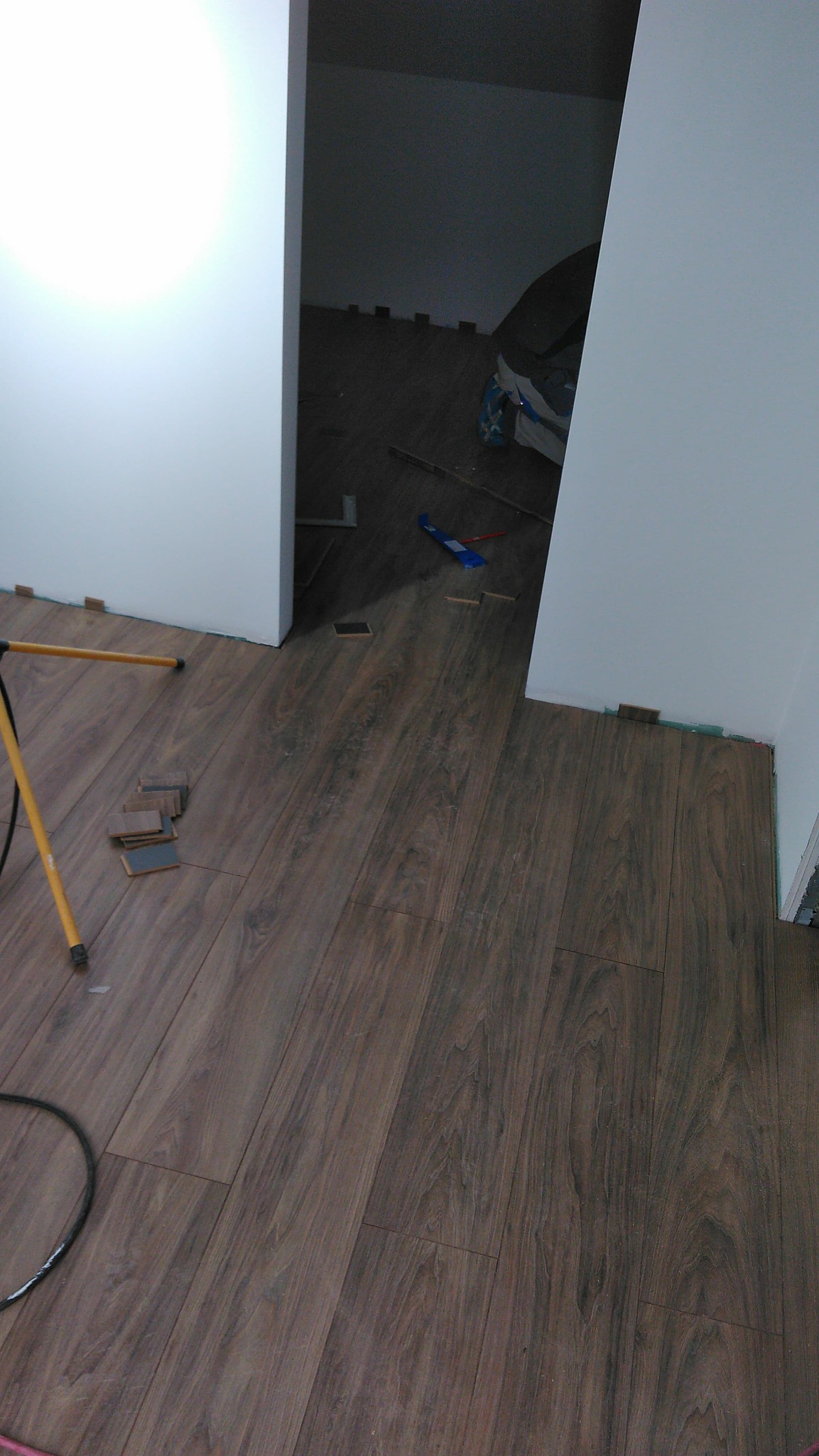 Układanie paneli podłogowych o ciemnym, drewnopodobnym wzorze, widoczne kliny dystansowe przy ścianach, lampa robocza i narzędzia w tle, w trakcie prac wykończeniowych w pomieszczeniu.