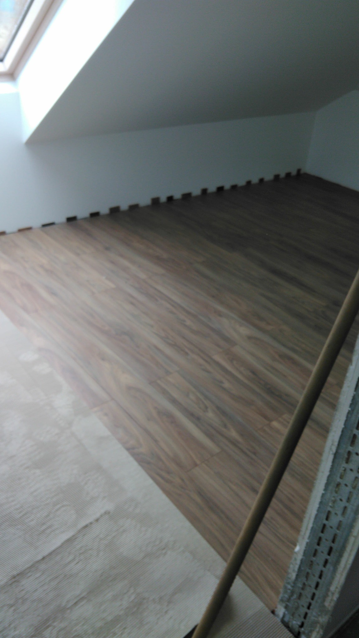 Fragment poddasza z częściowo ułożoną podłogą z paneli imitujących drewno, widoczna przestrzeń pod skosem dachu i okno dachowe.