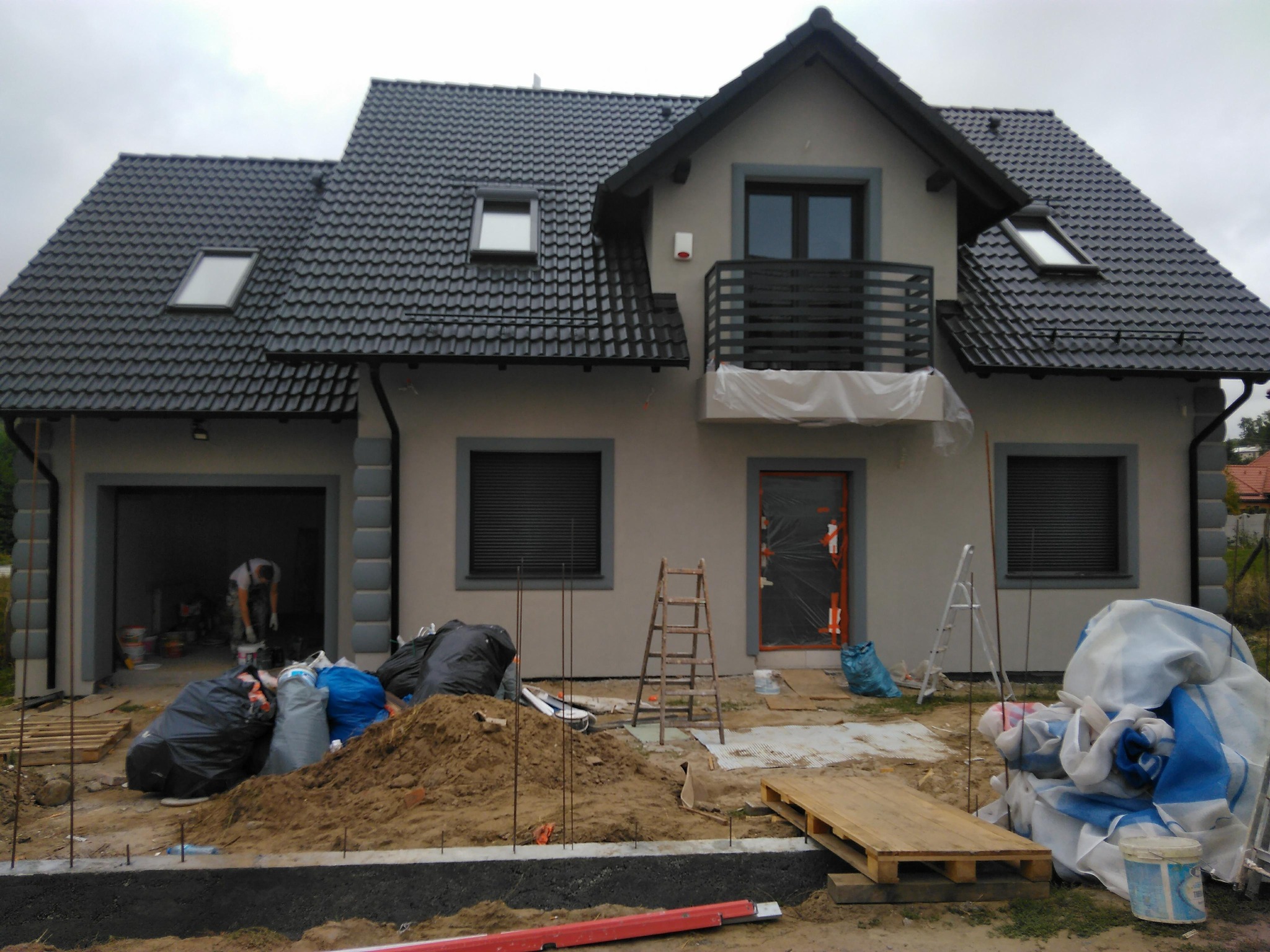Szary dom z ciemną dachówką w trakcie prac wykończeniowych. Widoczne worki z odpadami, ziemia i narzędzia. Okna i drzwi zabezpieczone folią.