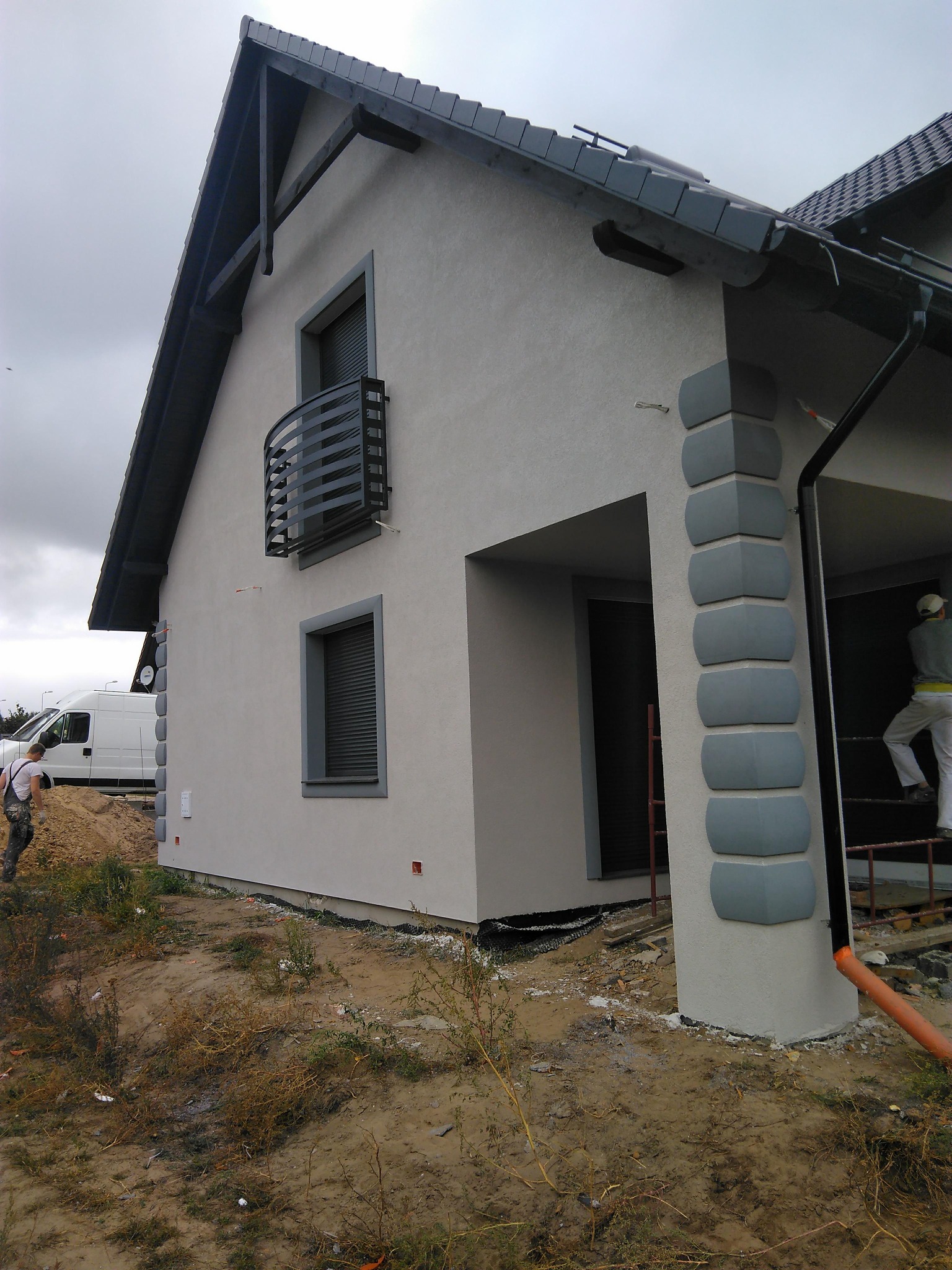 Elewacja domu jednorodzinnego po malowaniu w Dychowie. Szare detale architektoniczne, widoczni pracownicy podczas prac wykończeniowych i biały bus w tle.