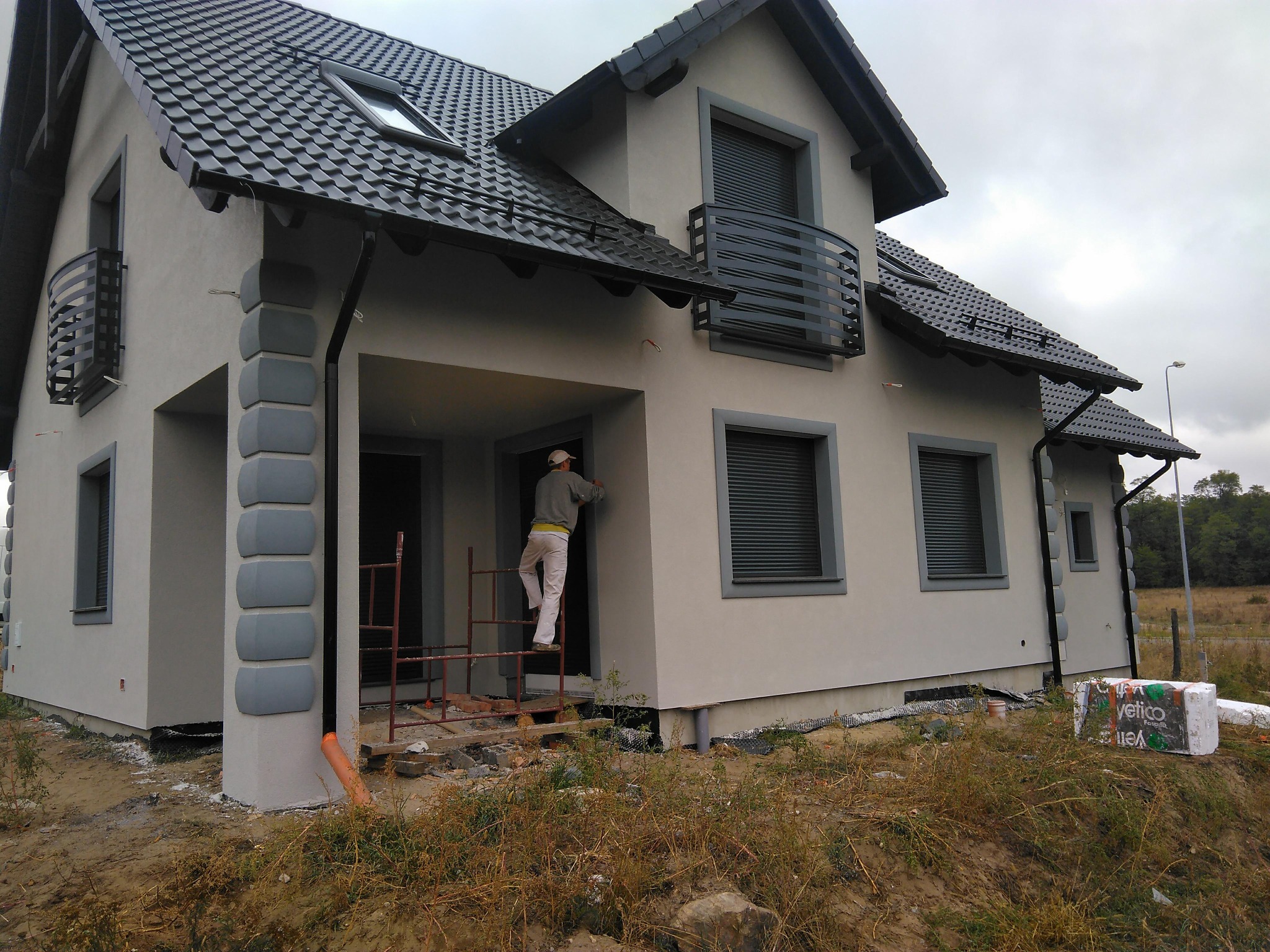 Ocieplanie elewacji domu jednorodzinnego. Pracownik na rusztowaniu aplikuje tynk. Widoczne okna z roletami zewnętrznymi, fragment dachu i ocieplenie.