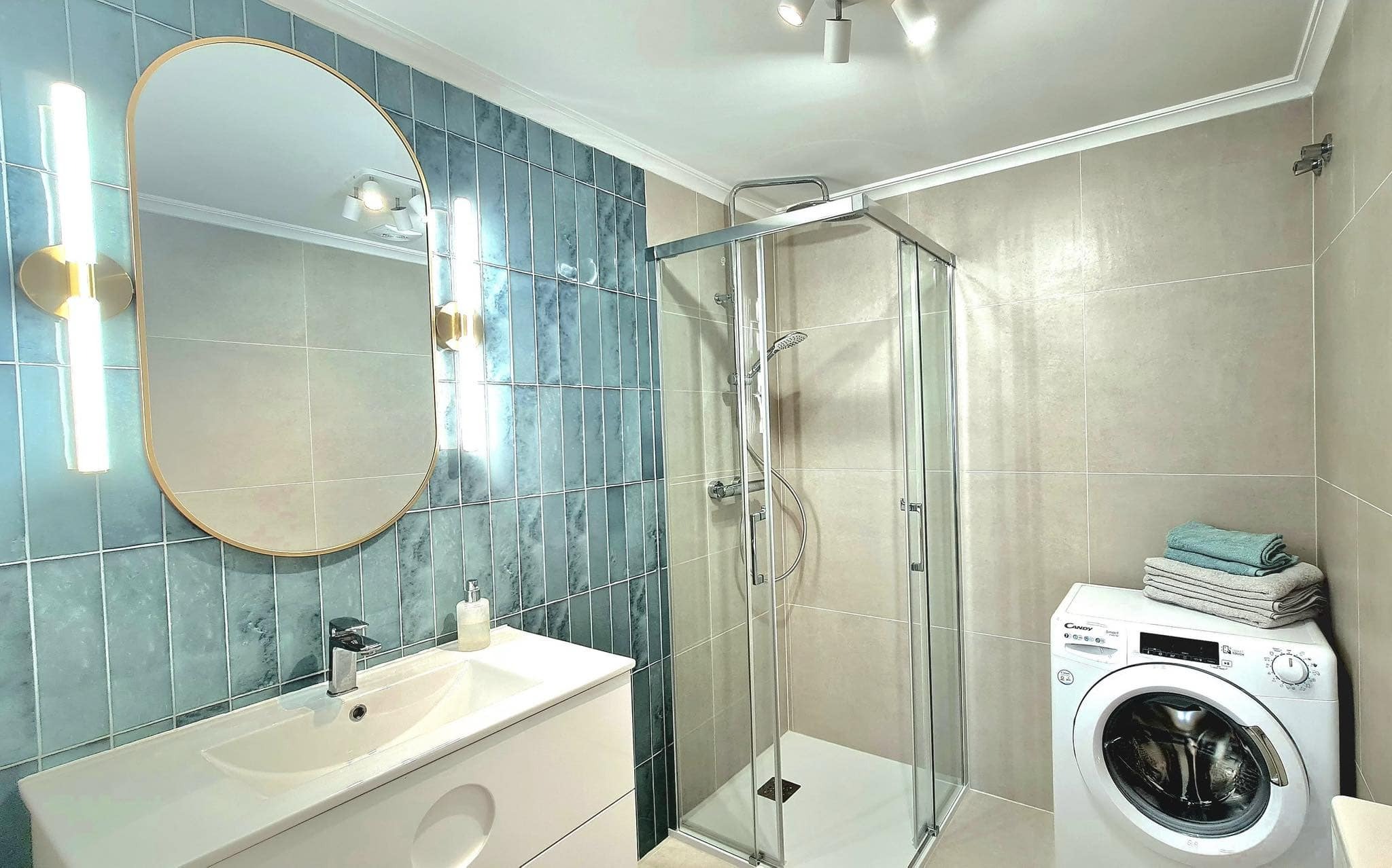 Nowoczesna łazienka z kabiną prysznicową, umywalką z szafką, owalnym lustrem z oświetleniem LED i pralką Candy z ułożonymi ręcznikami.