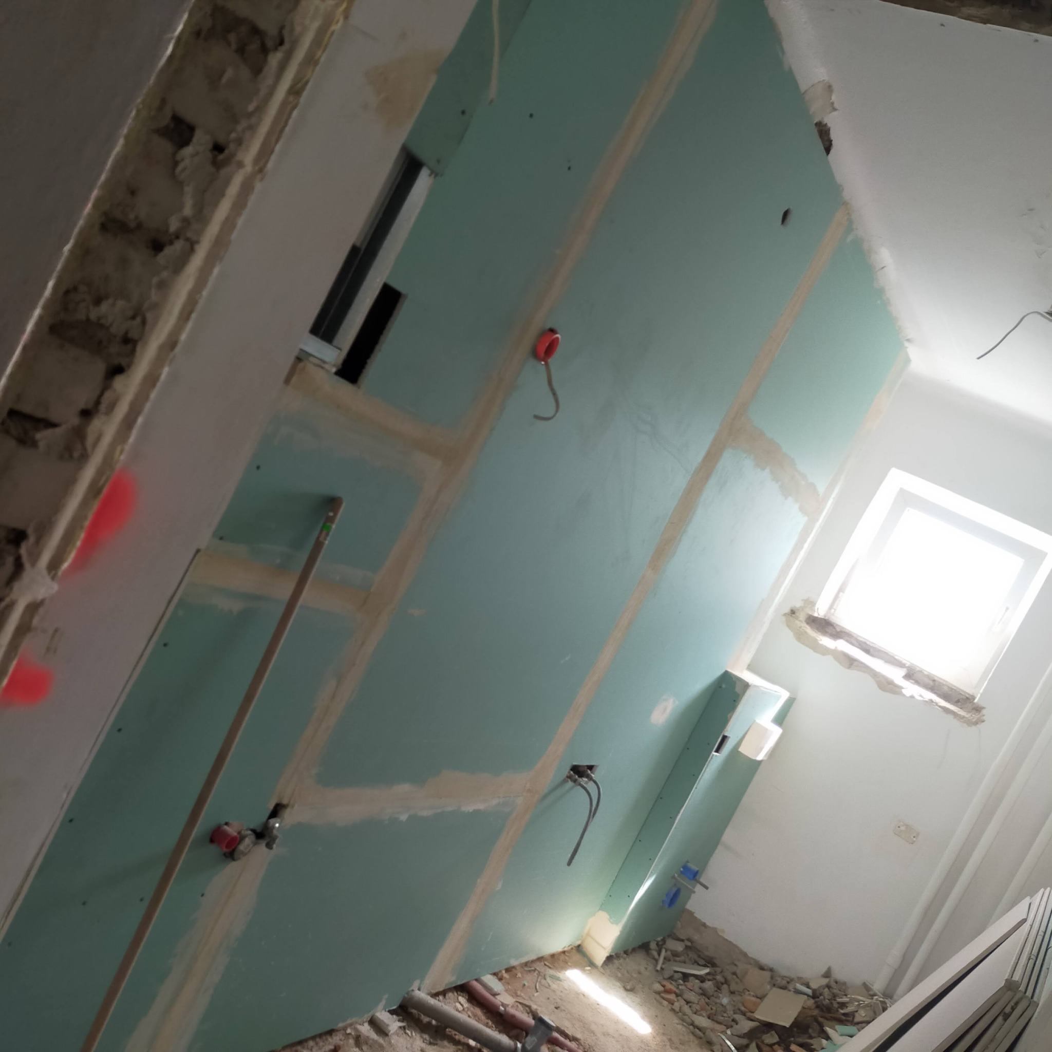 Montaż płyt kartonowo-gipsowych na ścianach w trakcie prac wykończeniowych, widoczne łączenia płyt wzmocnione taśmą, instalacja elektryczna wyprowadzona ze ściany, gruz na podłodze.