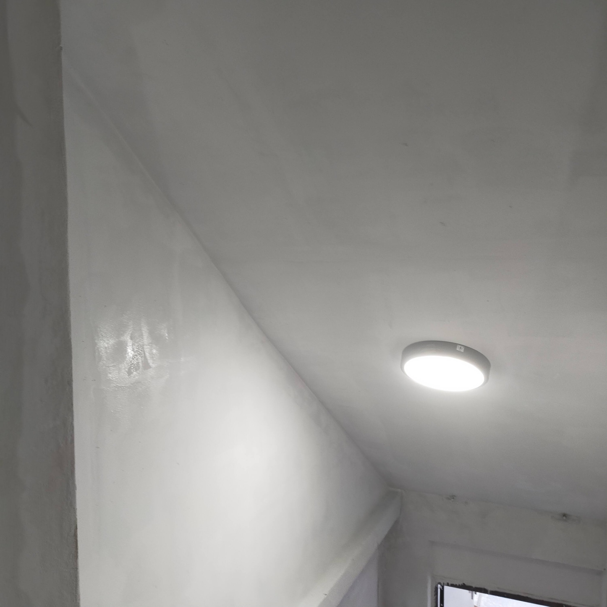 Białe, świeżo wyszpachlowane ściany i sufit pod skosem, oświetlone okrągłą lampą LED, widoczne ślady po szpachlowaniu w narożniku.