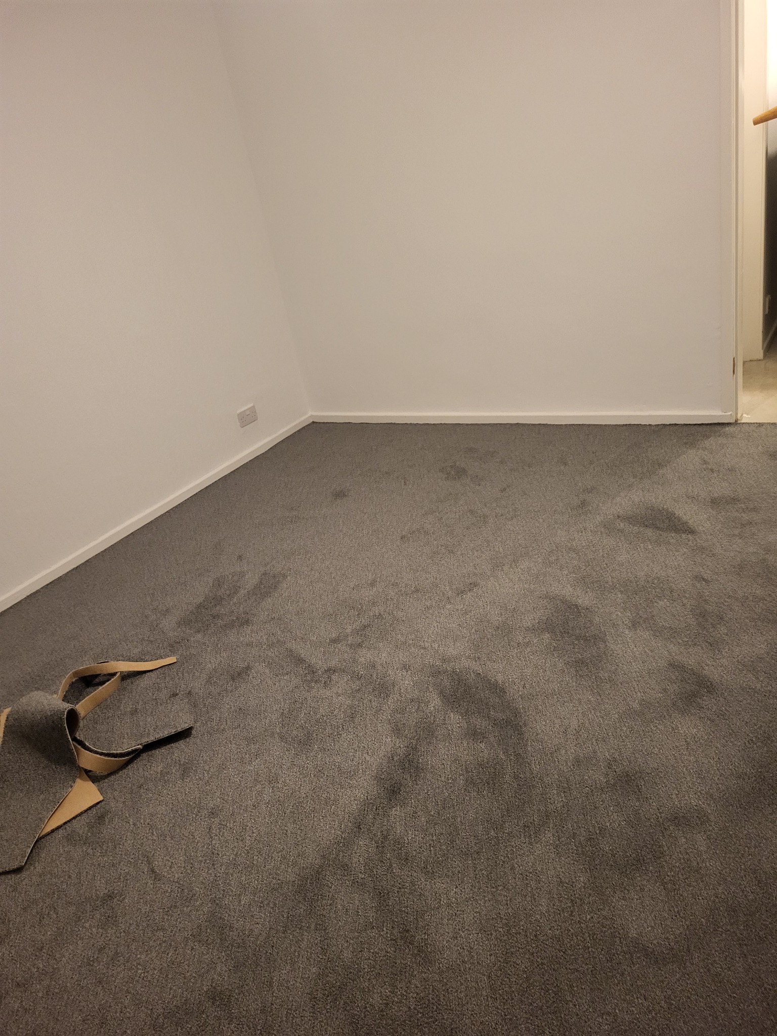 Pokój z nową, szarą wykładziną dywanową. Na podłodze leżą resztki wykładziny z widocznym brązowym spodem. Ściany pomalowane na biało.
