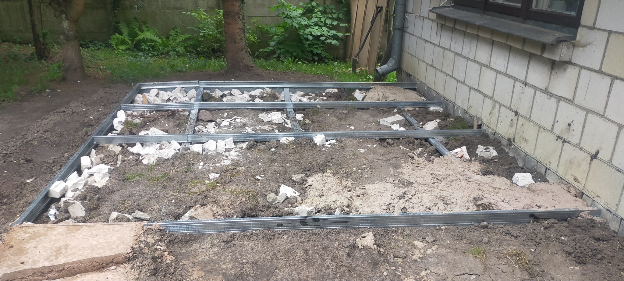Metalowa konstrukcja szkieletowa tarasu na ziemi, z fragmentami gruzu i piasku, przy ścianie budynku z białej cegły.
