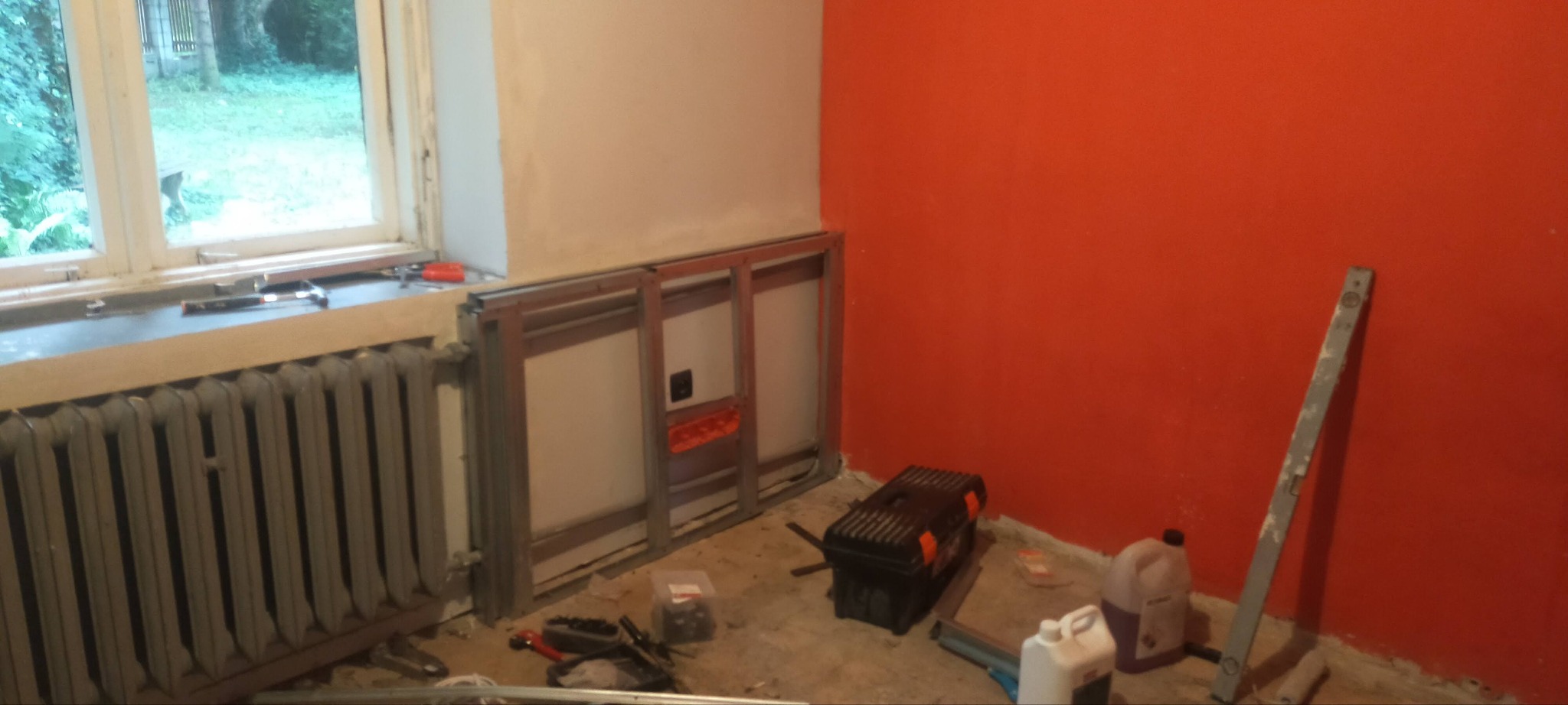Metalowy stelaż pod płytę kartonowo-gipsową na ścianie, widoczny stary grzejnik pod oknem, narzędzia i chemia budowlana na podłodze, pomarańczowa ściana w tle.