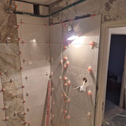 G-N.gut - Ściana w trakcie układania płytek, z widocznymi krzyżykami dystansowymi i poziomicą, oświetlona lampą roboczą. Fragment widocznej instalacji elektrycznej.