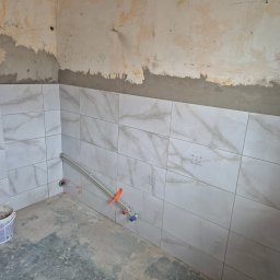 G-N.gut - Łazienka w trakcie remontu: białe płytki z marmurowym wzorem na ścianach, widoczne rury instalacji wodnej. Ściany częściowo przygotowane do wykończenia.