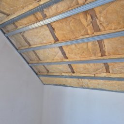 G-N.gut - Sufit podwieszany z izolacją: metalowa konstrukcja, wełna mineralna, białe ściany. Kątowe ujęcie z dołu, widoczne mocowania i detale izolacji.