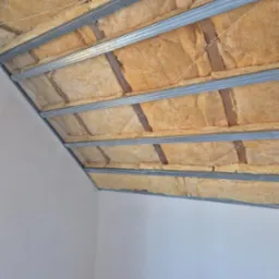 Sufit podwieszany z izolacją: metalowa konstrukcja, wełna mineralna, białe ściany. Kątowe ujęcie z dołu, widoczne mocowania i detale izolacji.