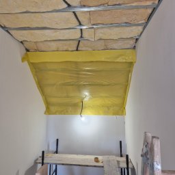G-N.gut - Izolacja stropu wełną mineralną, zabezpieczona folią żółtą, widoczna konstrukcja metalowa, rusztowanie i drabina w pomieszczeniu w trakcie prac wykończeniowych.