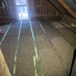 G-N.gut - Izolacja poddasza z widocznymi belkami stropowymi i oknem dachowym. Granulat izolacyjny równomiernie rozłożony między belkami. Prace wykończeniowe w toku.