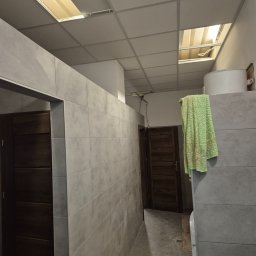 G-N.gut - Wykończona łazienka w Izdebkach z szarą glazurą, widoczne drzwi z ciemnego drewna i sufit podwieszany z oświetleniem. Na ścianie wisi zielony ręcznik.