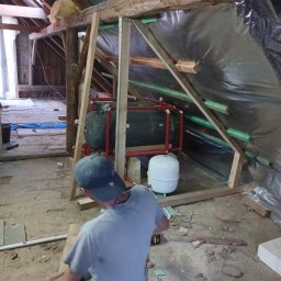 G-N.gut - Prace wykończeniowe na poddaszu: montaż instalacji grzewczej, izolacja folią, drewniana konstrukcja szkieletowa. Mężczyzna w czapce przy pracy z drewnem. Widoczne narzędzia.