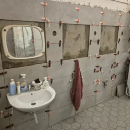 Remont łazienki w trakcie: szare płytki z krzyżykami dystansowymi na ścianach, umywalka z kosmetykami i lustro. Widoczny bojler i pralka. Częściowo położona podłoga.