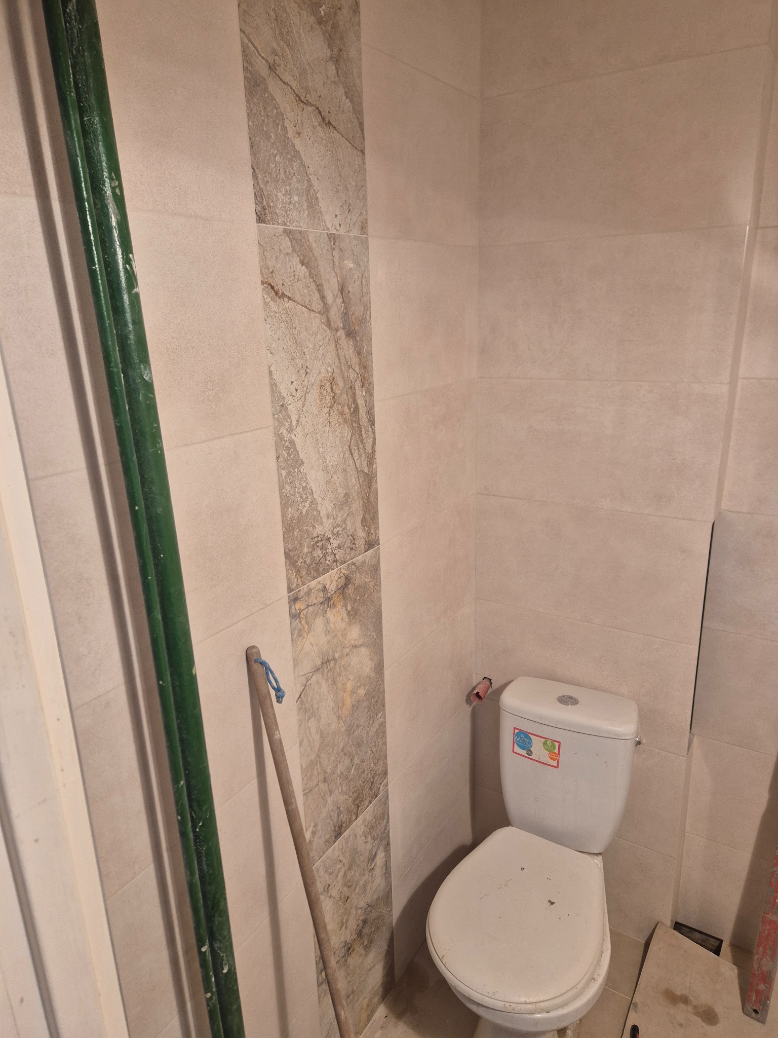 Wnętrze łazienki w trakcie prac: beżowe płytki imitujące kamień, fragment toalety, rury i narzędzia. Widoczne niedokończone detale.