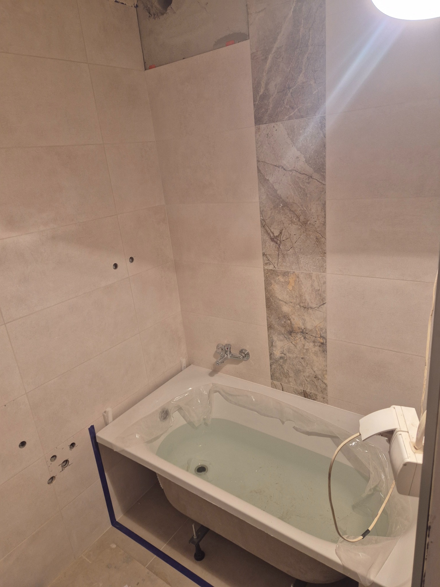 Wykończona łazienka z beżowymi płytkami, dekoracyjnym pasem z motywem marmuru i wanną zabezpieczoną folią. Widoczne otwory na armaturę.