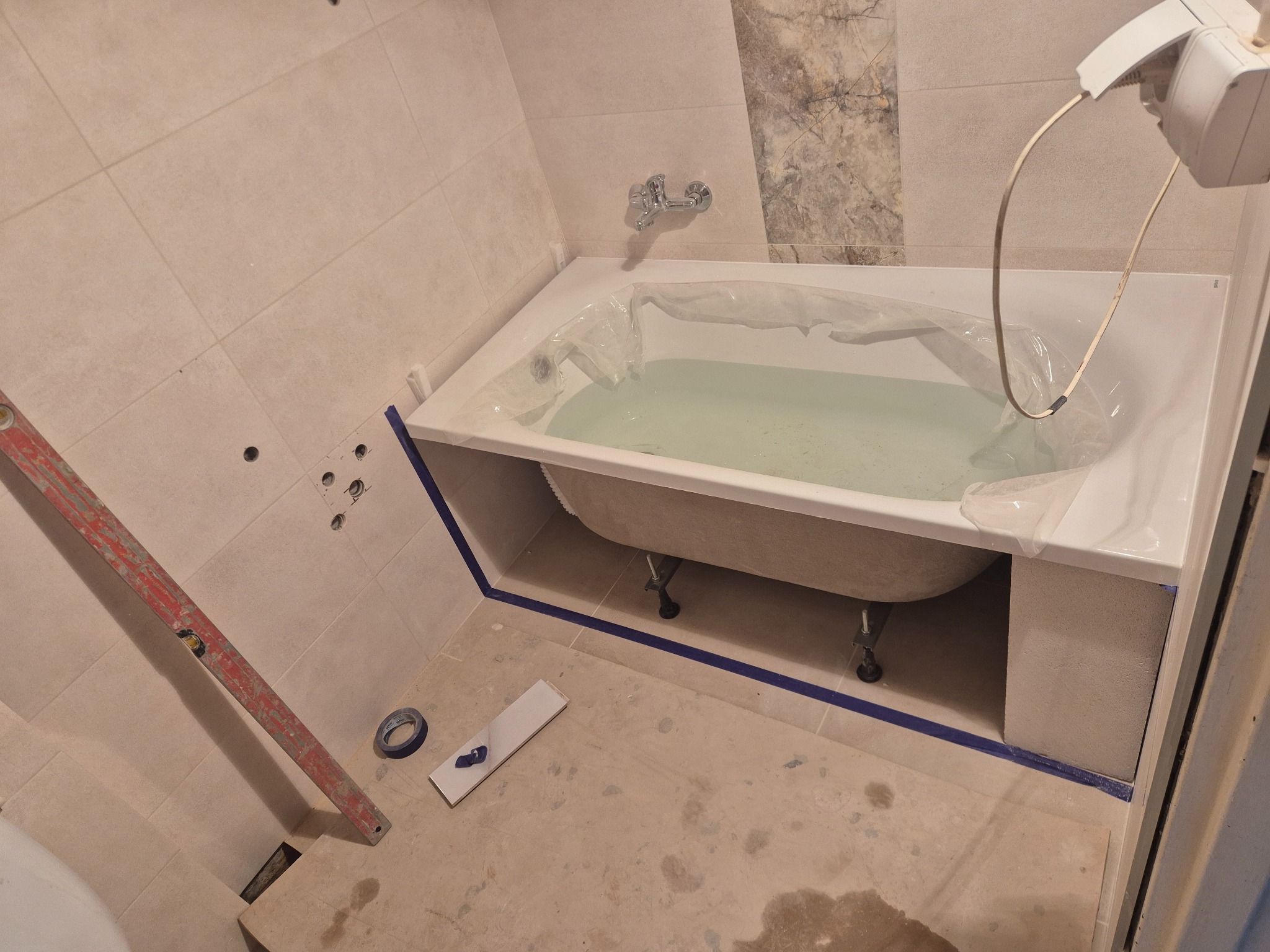 Przygotowania do remontu łazienki: wanna z wodą zabezpieczona folią, poziomica, taśma malarska, płytki. Widoczne otwory w ścianie i fragment dekoracyjnej płytki.