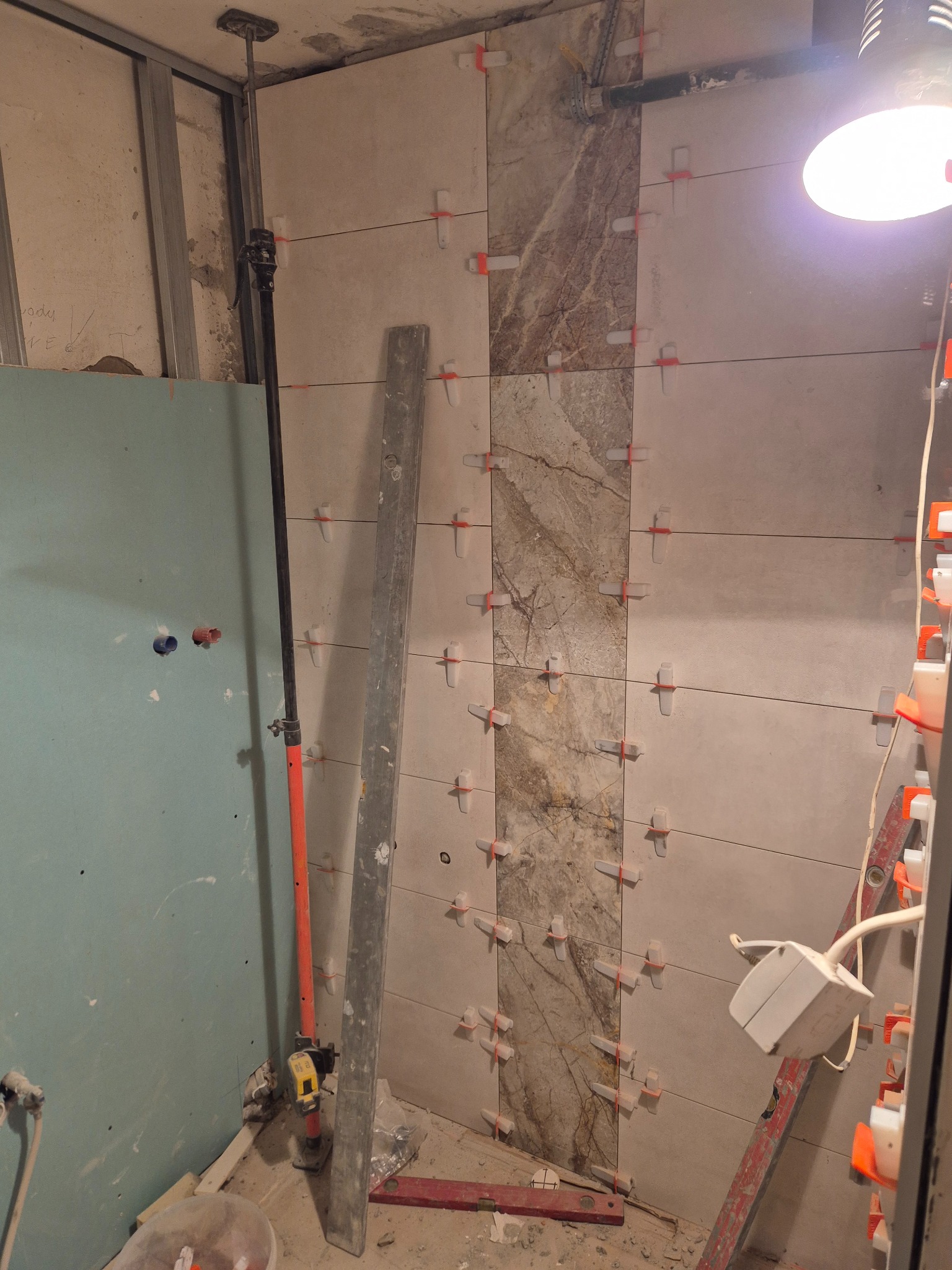 Ściana łazienki w trakcie wykańczania: ułożone płytki ceramiczne z krzyżykami dystansowymi, widoczna płyta karton-gips i narzędzia. Oświetlenie robocze.