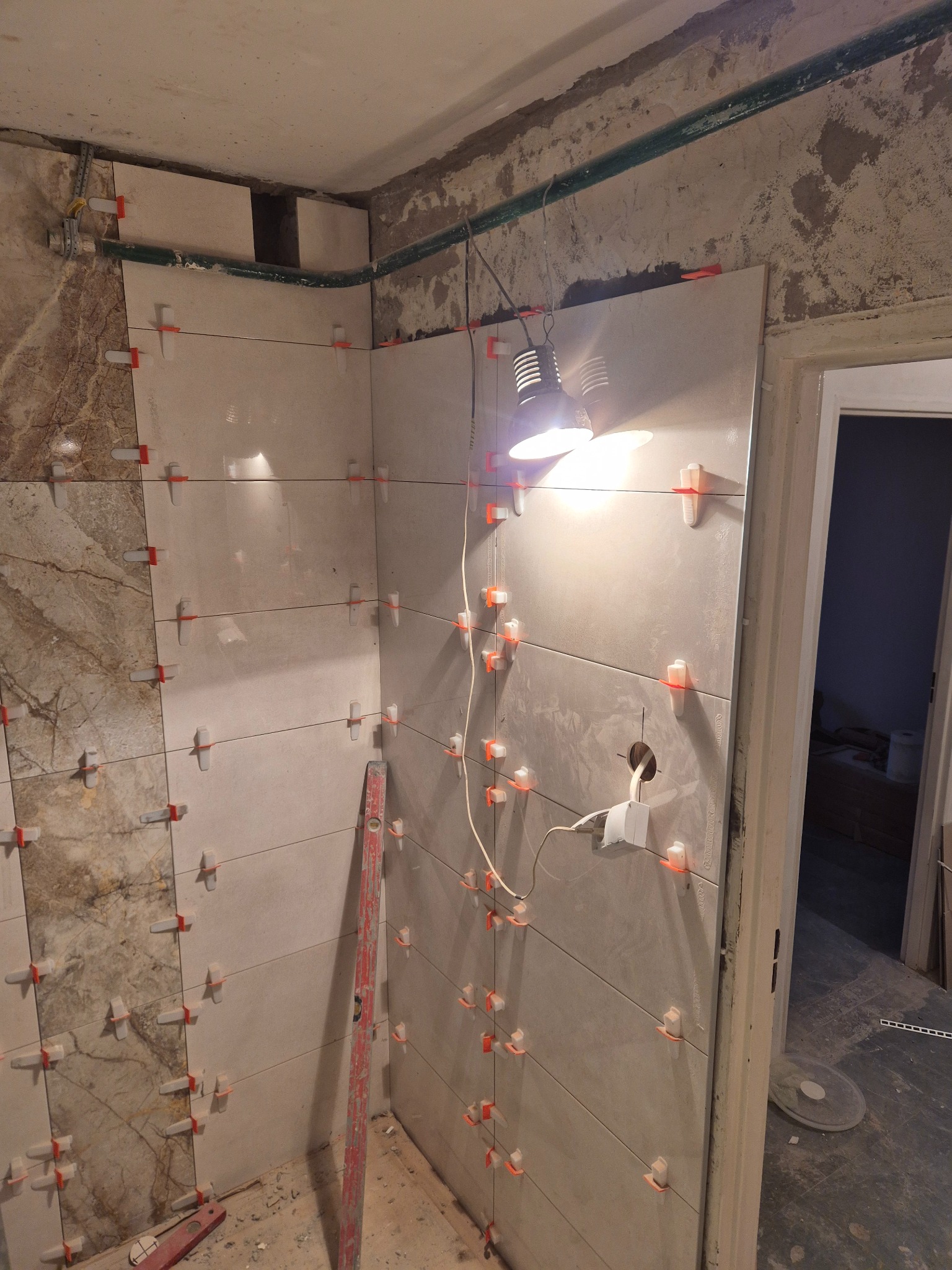 Ściana w trakcie układania płytek, z widocznymi krzyżykami dystansowymi i poziomicą, oświetlona lampą roboczą. Fragment widocznej instalacji elektrycznej.