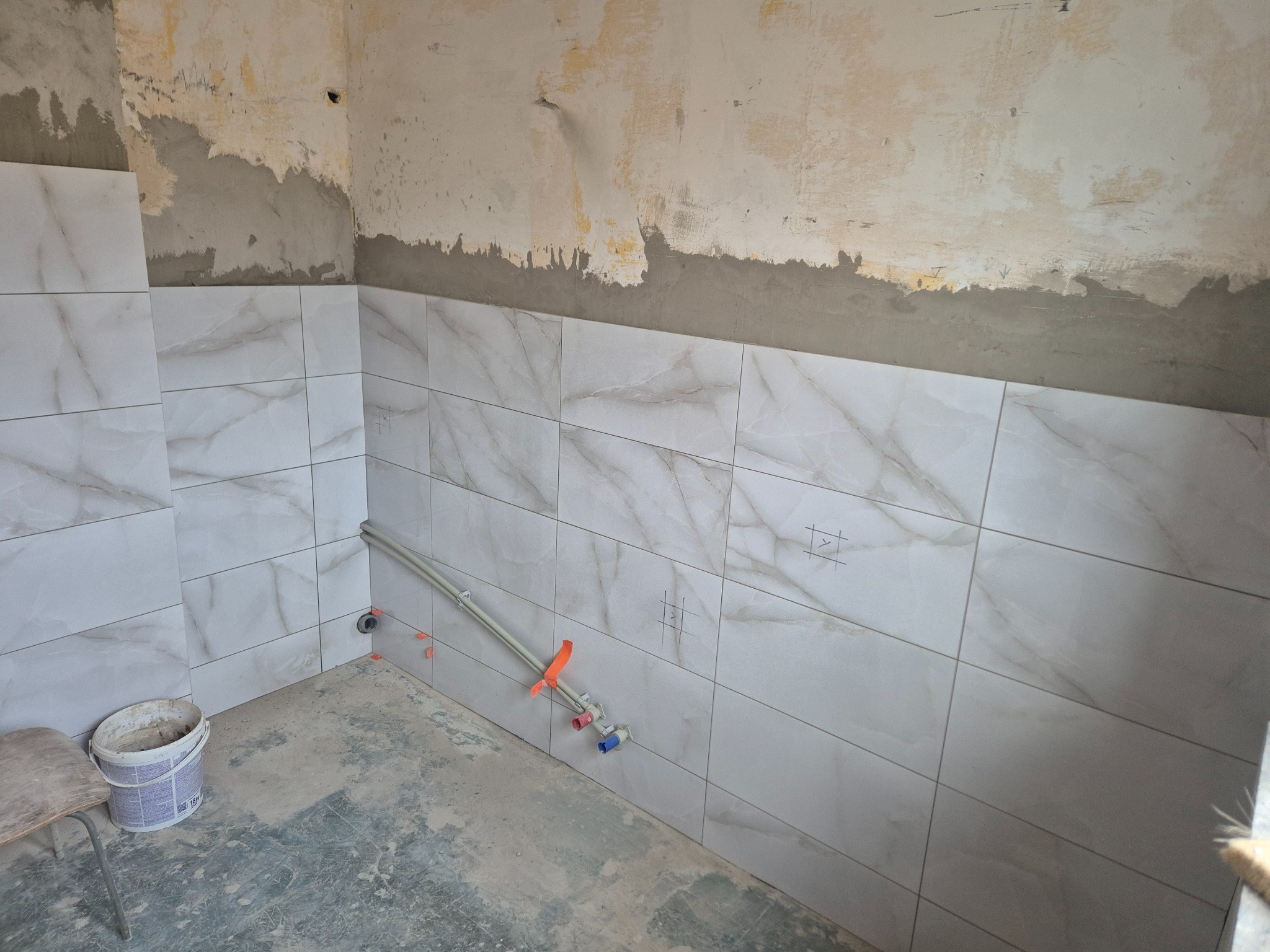 Łazienka w trakcie remontu: białe płytki z marmurowym wzorem na ścianach, widoczne rury instalacji wodnej. Ściany częściowo przygotowane do wykończenia.