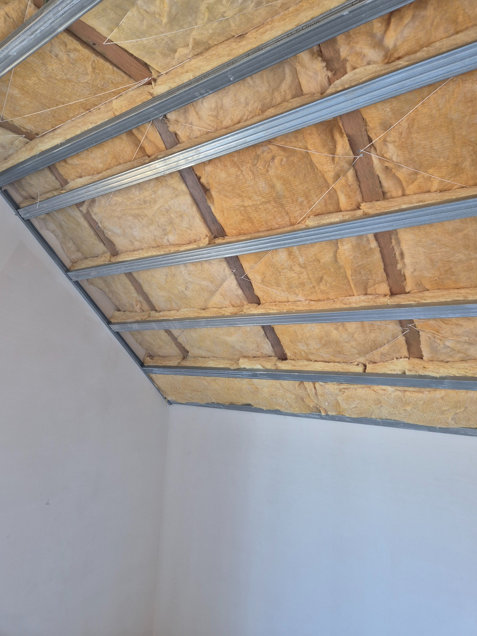 Sufit podwieszany z izolacją: metalowa konstrukcja, wełna mineralna, białe ściany. Kątowe ujęcie z dołu, widoczne mocowania i detale izolacji.