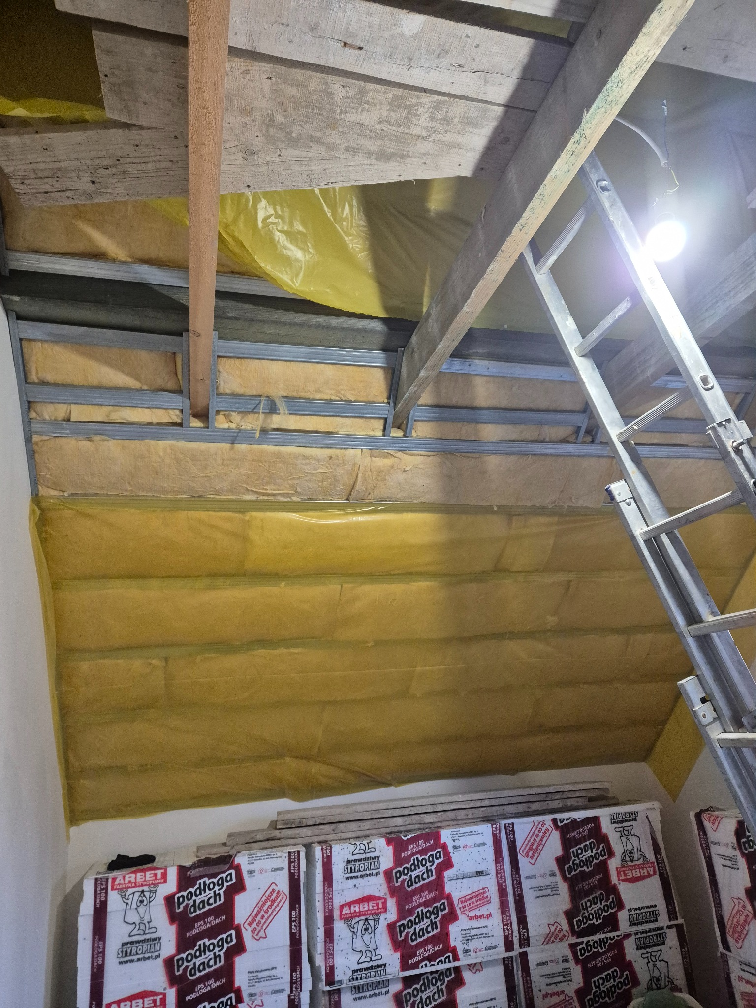 Ocieplenie poddasza wełną mineralną, widoczne metalowe profile, folia paroizolacyjna i opakowania styropianu. Prace wykończeniowe na poddaszu.
