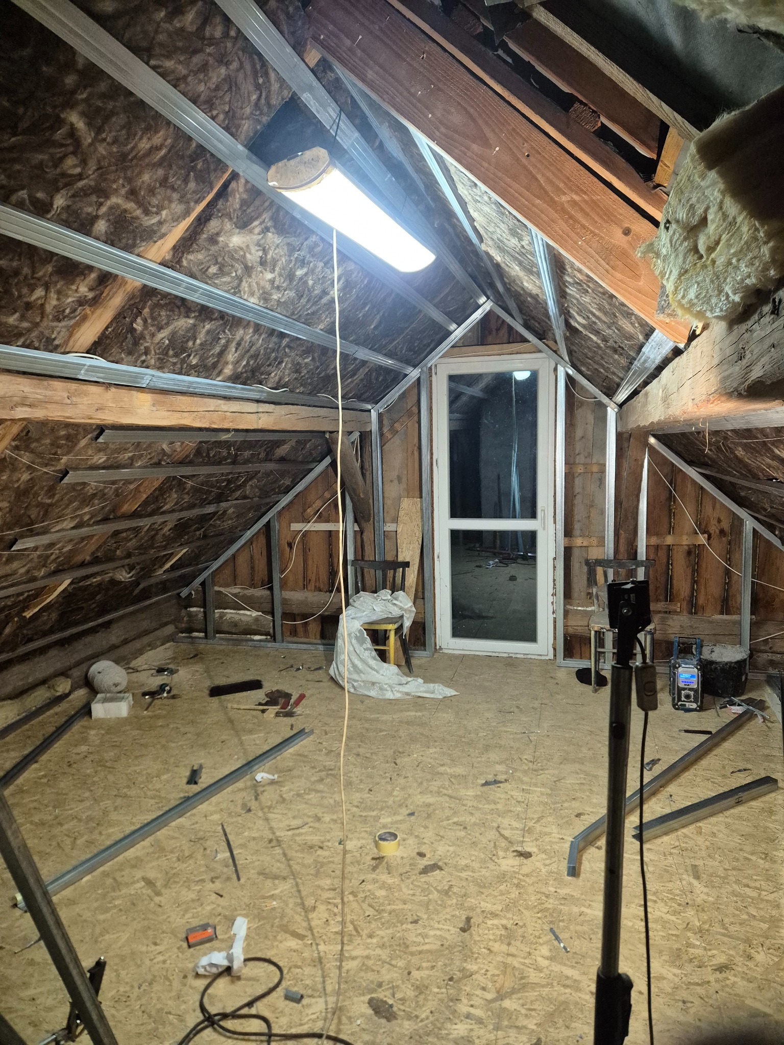 Adaptacja poddasza: metalowy szkielet pod zabudowę, widoczne ocieplenie, okno dachowe i narzędzia na podłodze. Prace w toku, surowe wykończenie.