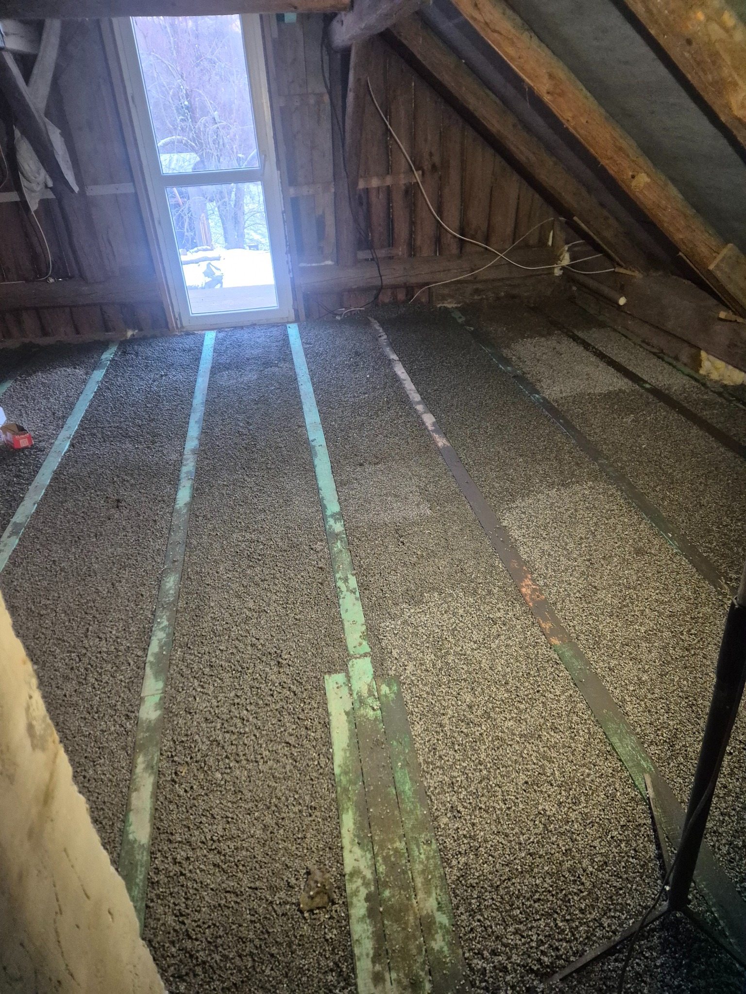 Izolacja poddasza z widocznymi belkami stropowymi i oknem dachowym. Granulat izolacyjny równomiernie rozłożony między belkami. Prace wykończeniowe w toku.