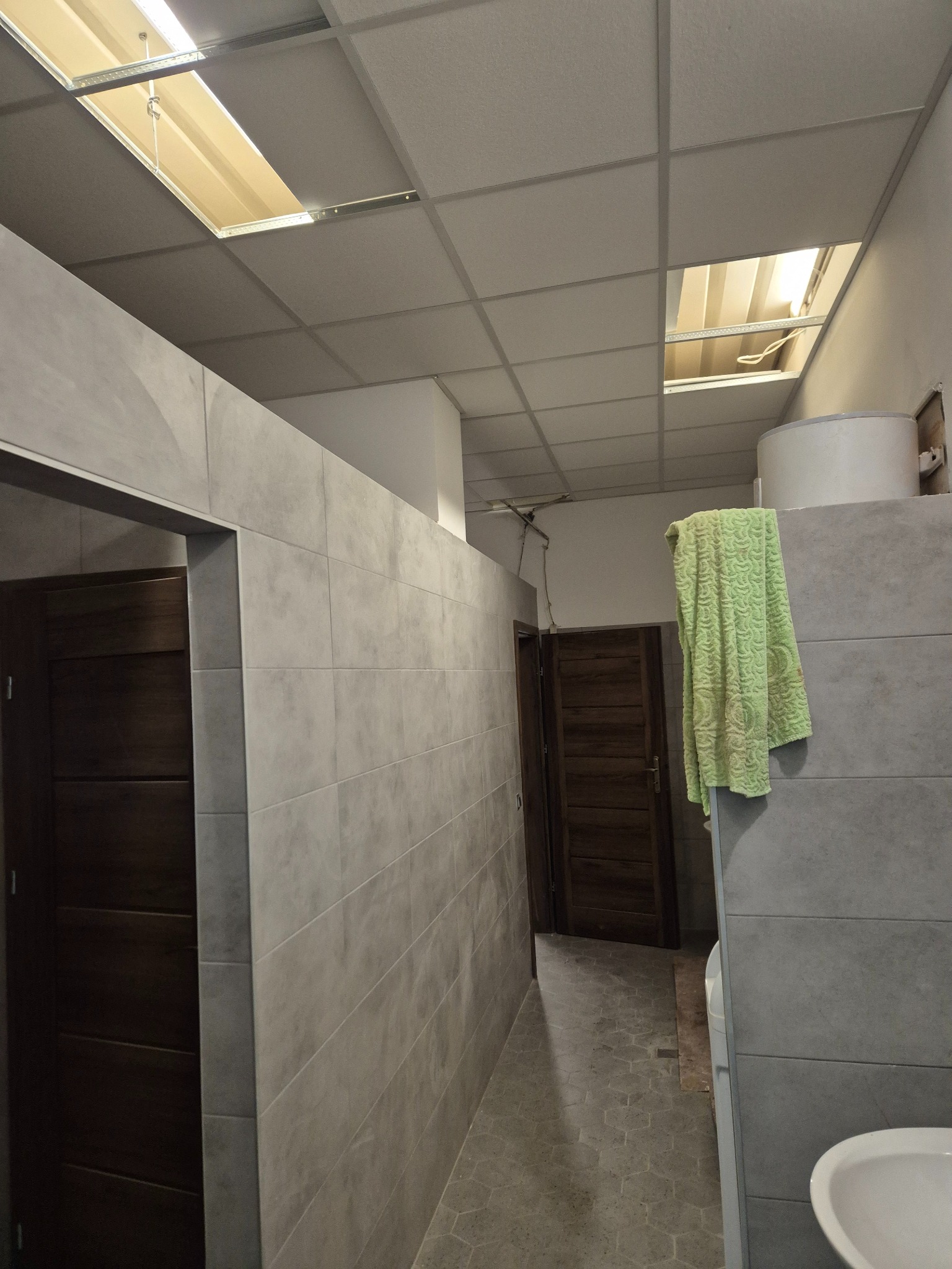 Wykończona łazienka w Izdebkach z szarą glazurą, widoczne drzwi z ciemnego drewna i sufit podwieszany z oświetleniem. Na ścianie wisi zielony ręcznik.