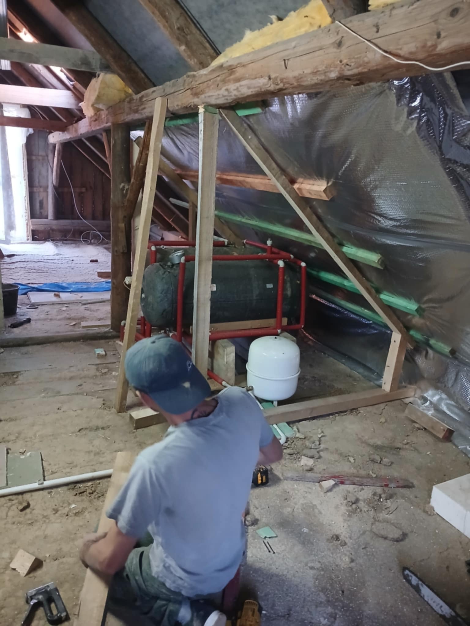 Prace wykończeniowe na poddaszu: montaż instalacji grzewczej, izolacja folią, drewniana konstrukcja szkieletowa. Mężczyzna w czapce przy pracy z drewnem. Widoczne narzędzia.