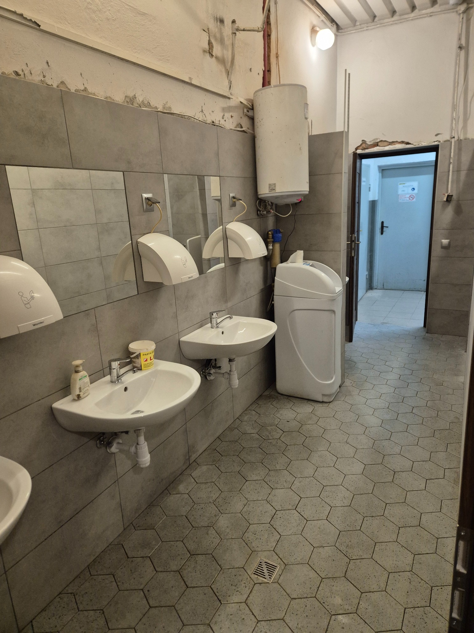 Szara łazienka z umywalkami, suszarkami do rąk i bojlerem. Na ścianach szare płytki. Podłoga wyłożona heksagonalnymi płytkami. Widoczny filtr do wody i otwarte drzwi.