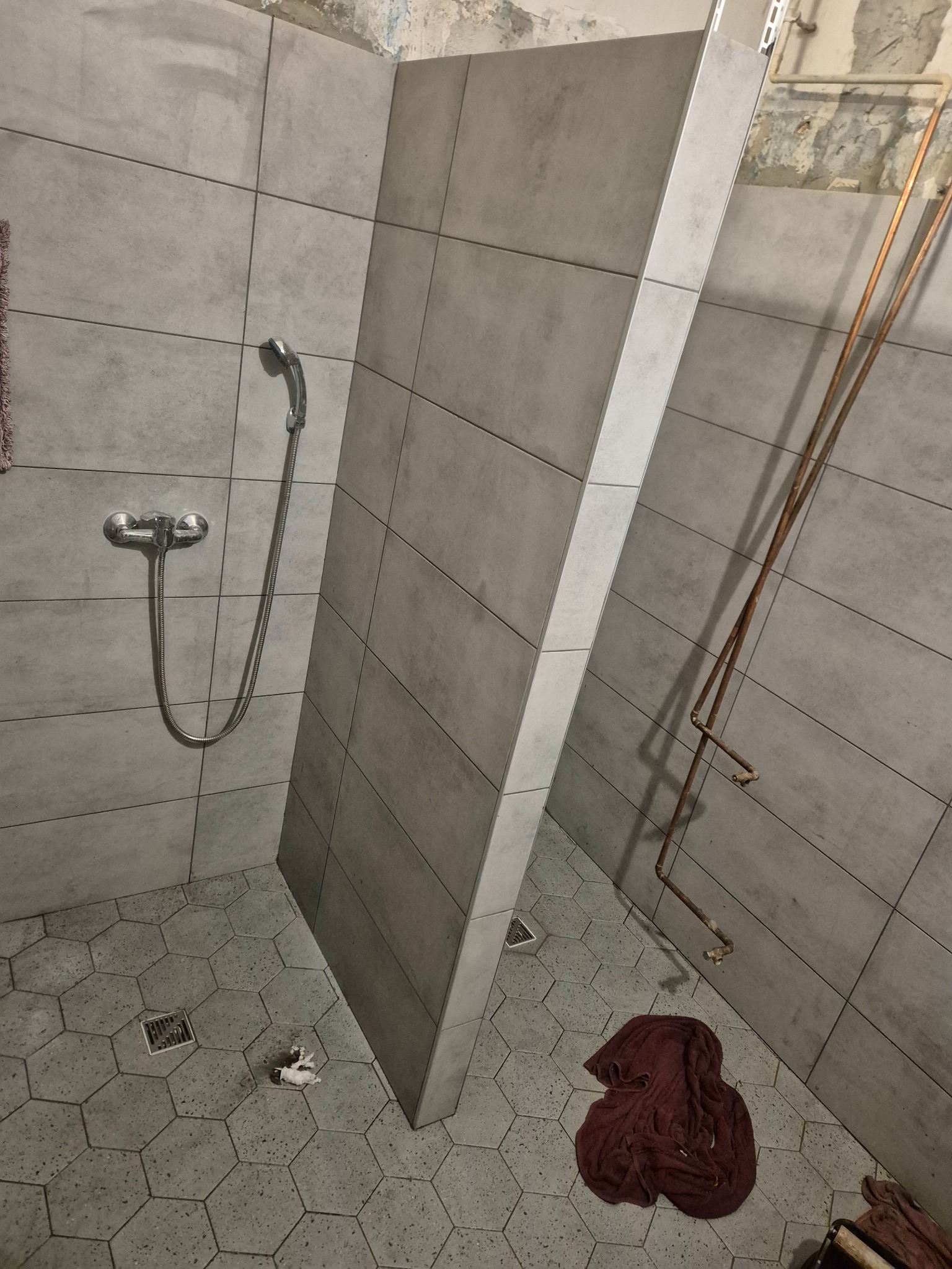 Szara łazienka w trakcie remontu: heksagonalne płytki podłogowe, prysznic z baterią, fragment ściany bez wykończenia, rury. Widoczne ślady prac remontowych.