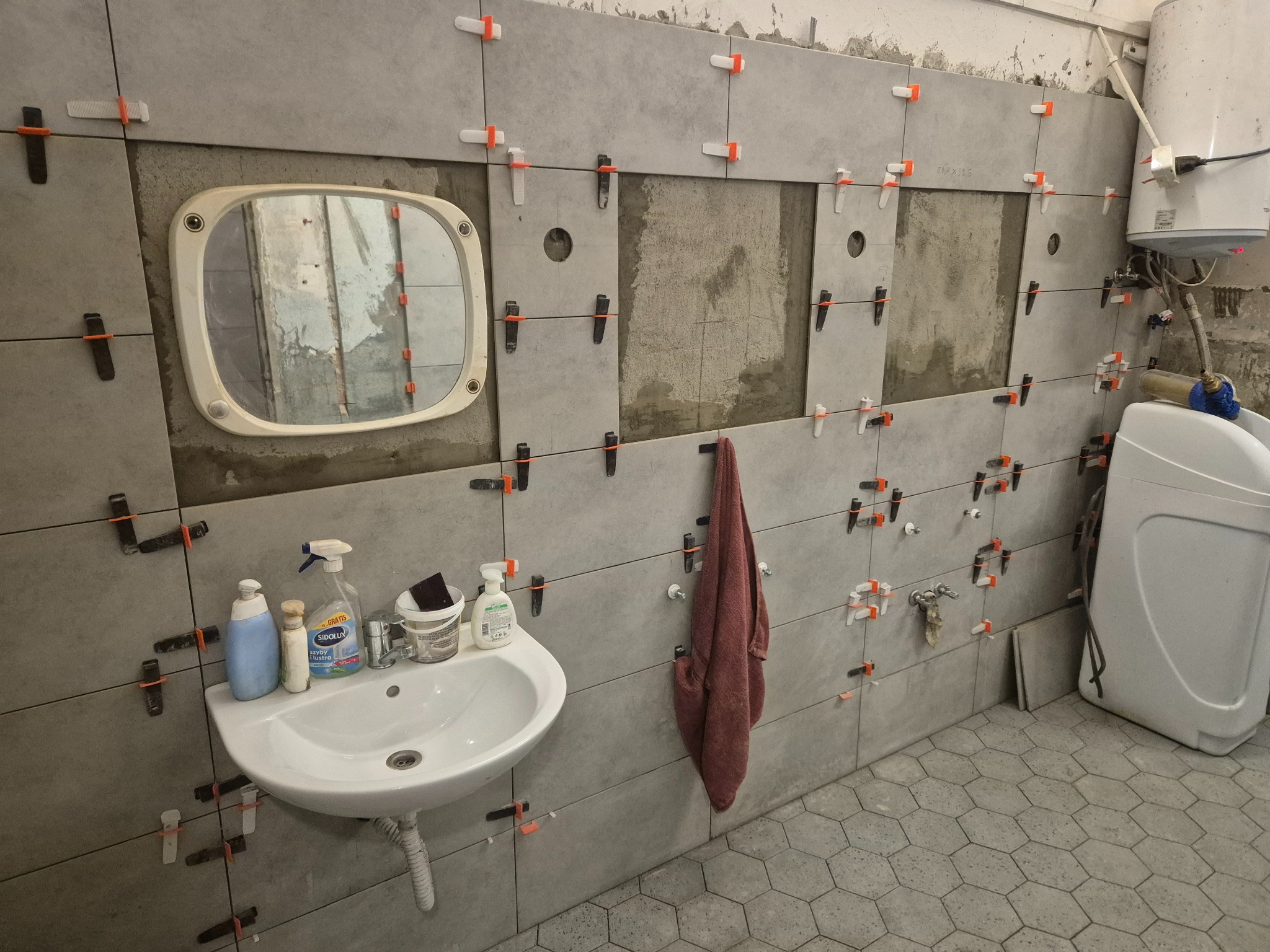 Remont łazienki w trakcie: szare płytki z krzyżykami dystansowymi na ścianach, umywalka z kosmetykami i lustro. Widoczny bojler i pralka. Częściowo położona podłoga.