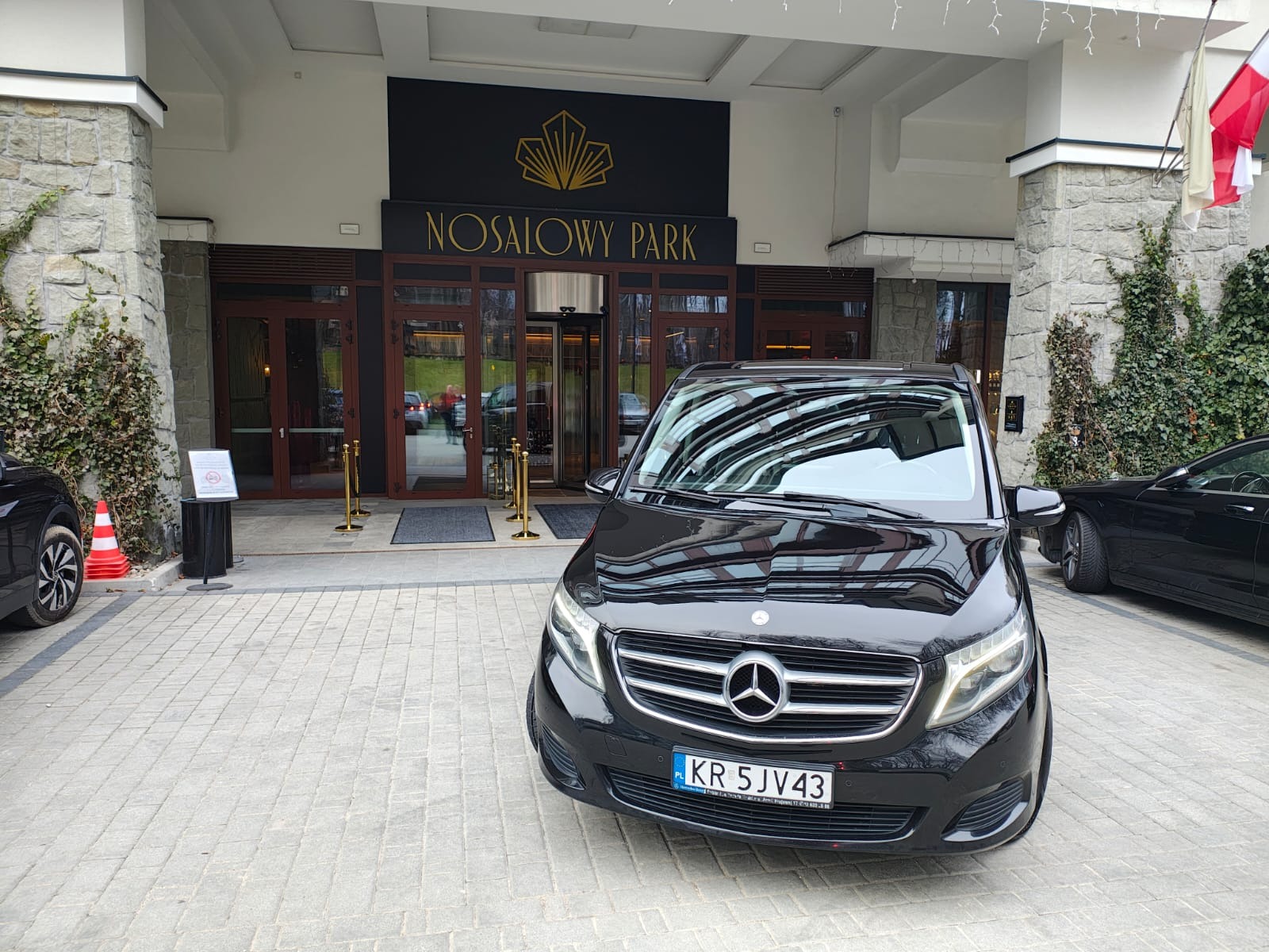 Czarny Mercedes V-klasy z numerem rejestracyjnym KR 5JV43 przed wejściem do Nosalowy Park w Zakopanem. Widoczne flagi Polski i hotelu.