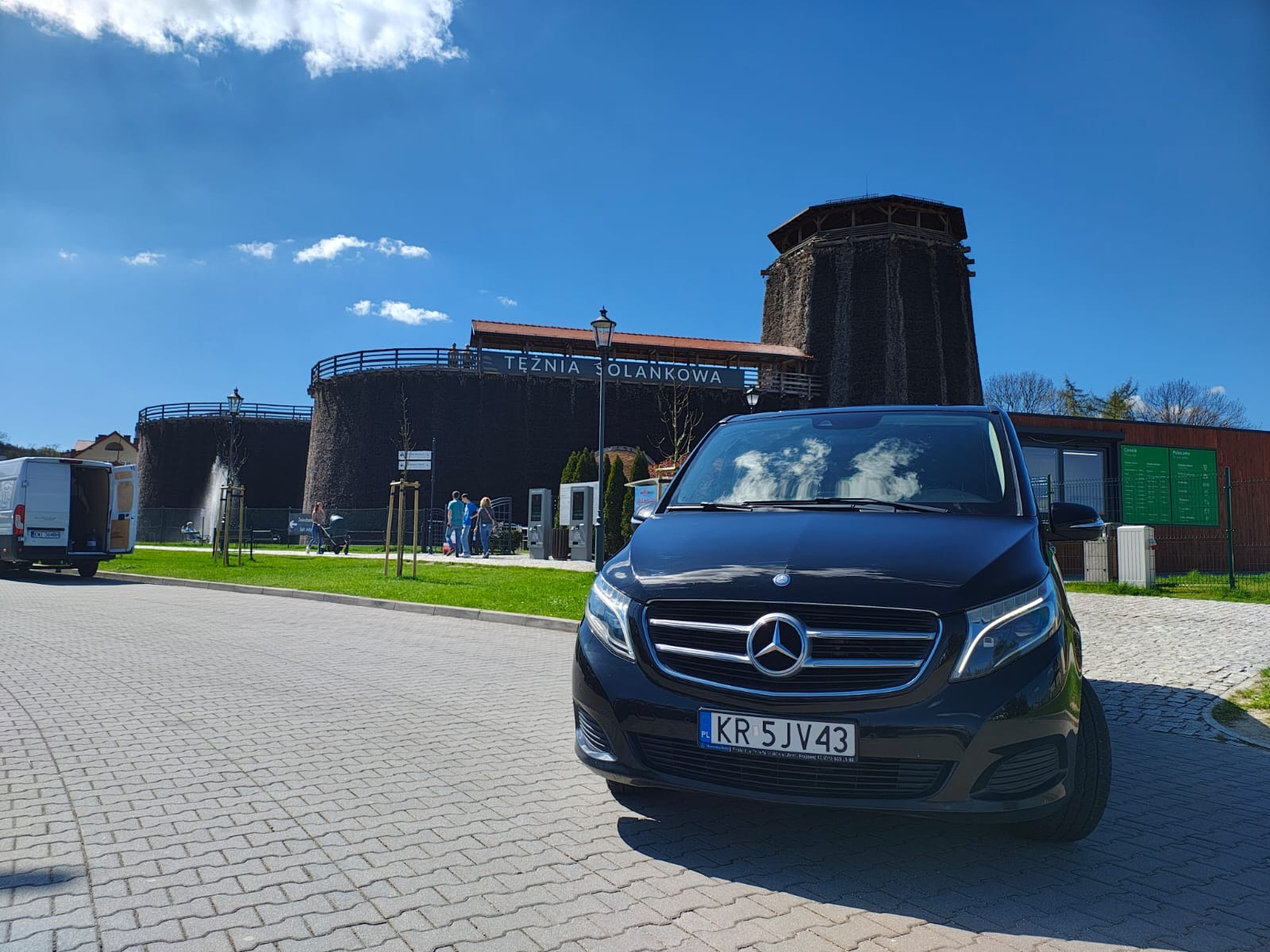 Czarny Mercedes V-klasa na tle Tężni Solankowej w Inowrocławiu. Błękitne niebo z chmurami odbija się w szybie samochodu. Świeci słońce, widoczni spacerujący ludzie.