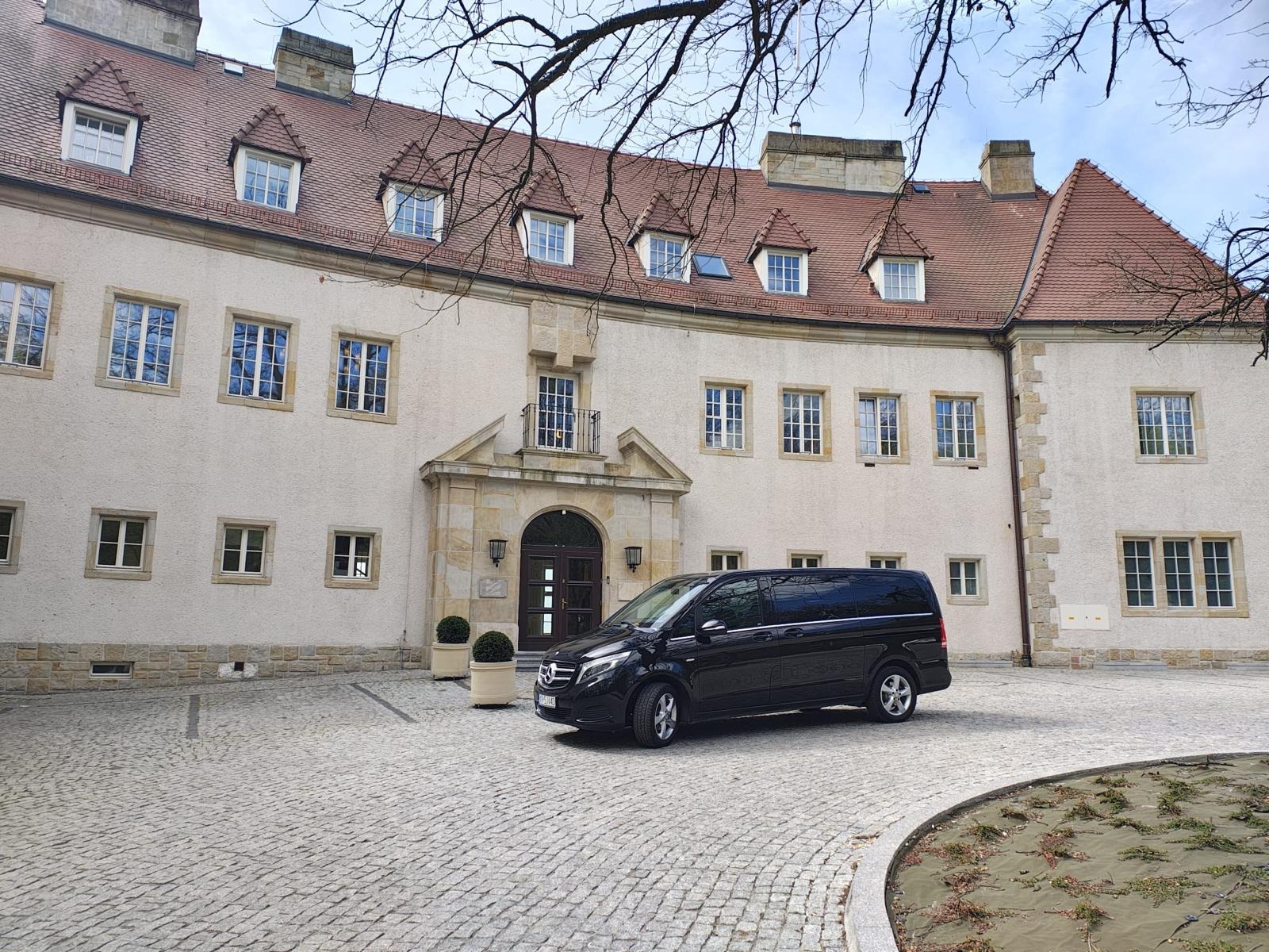 Czarny van Mercedes-Benz V-Klasa zaparkowany przed budynkiem z jasną fasadą i czerwonym dachem. Widoczne drzewa bez liści i kostka brukowa na podjeździe.