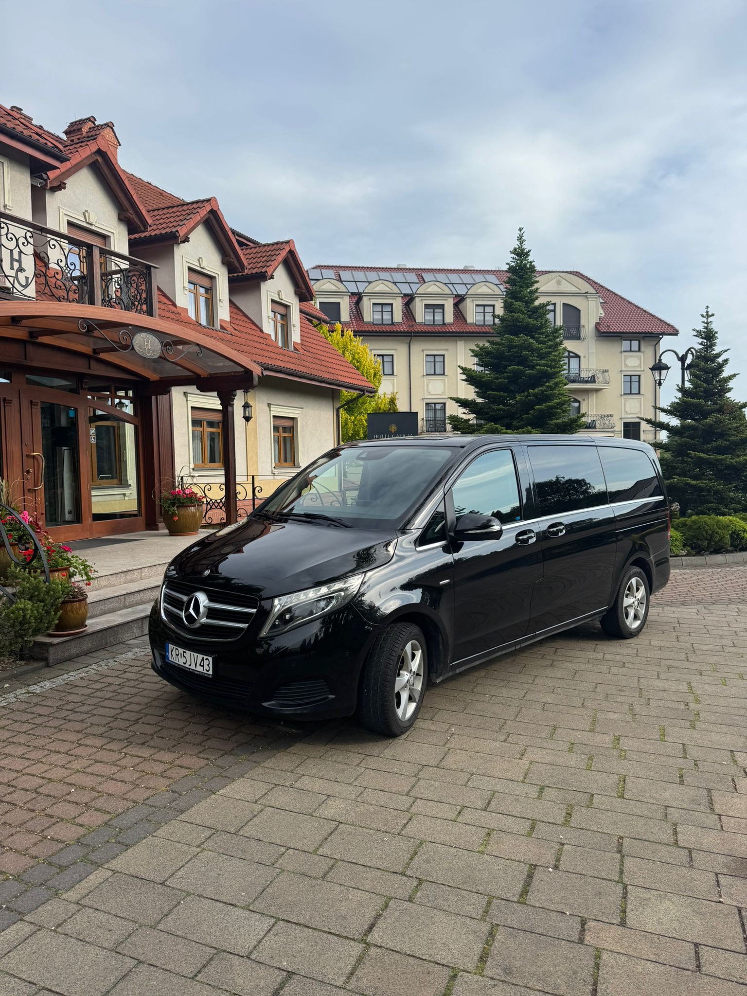 Czarny van Mercedes zaparkowany przed budynkiem z czerwonym dachem i zielonymi drzewami. Tablica rejestracyjna widoczna. Usługi transportowe.